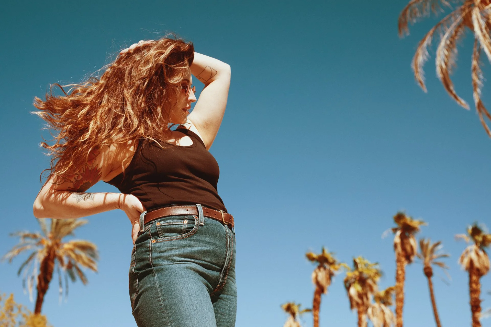 A woman with curly red hair wearing a black tank top and blue jeans, standing outdoors under a clear blue sky with palm trees in the background.