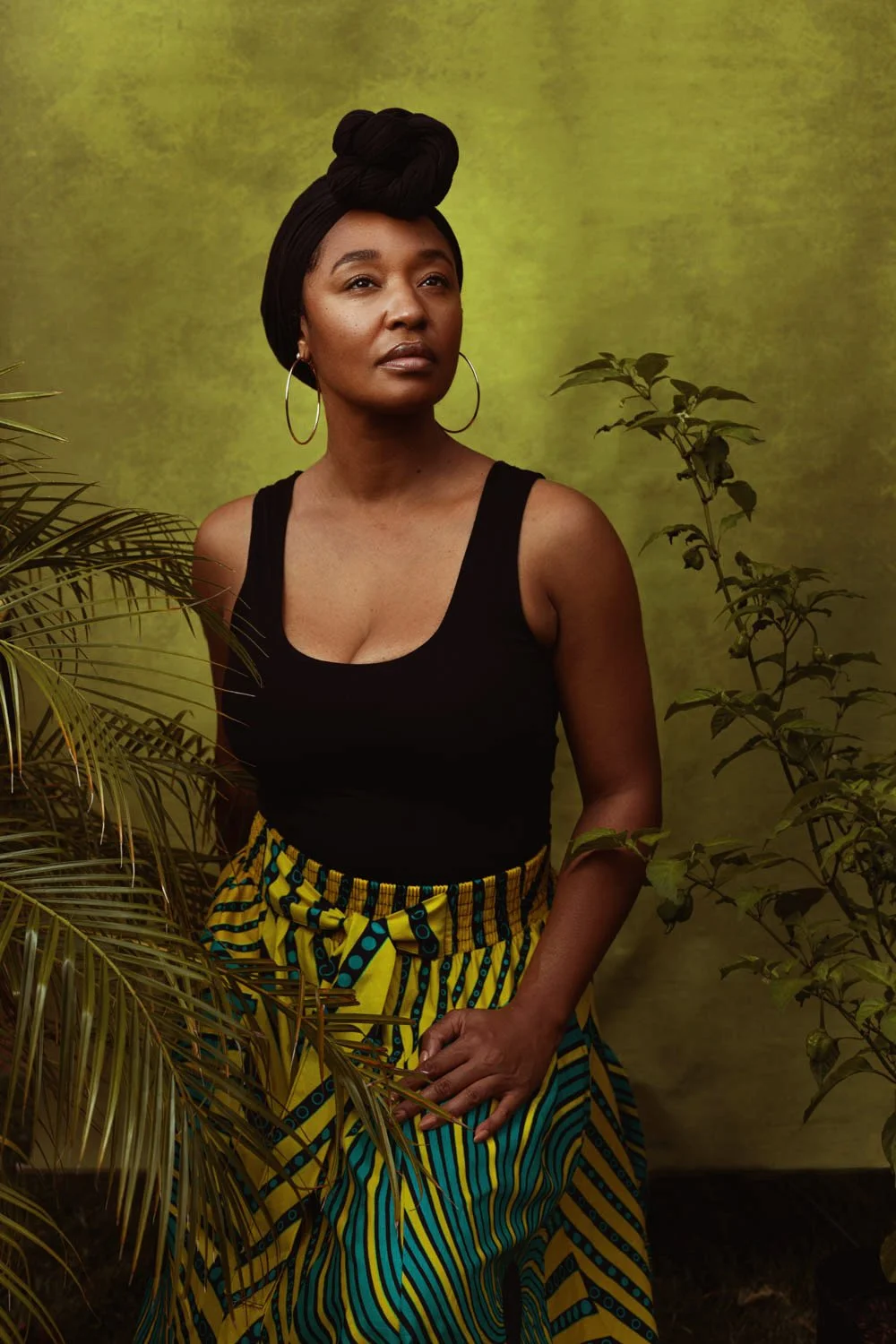 A woman with dark skin, wearing hoop earrings, a black sleeveless top, and a colorful patterned skirt, standing before a green textured background with plants around her.