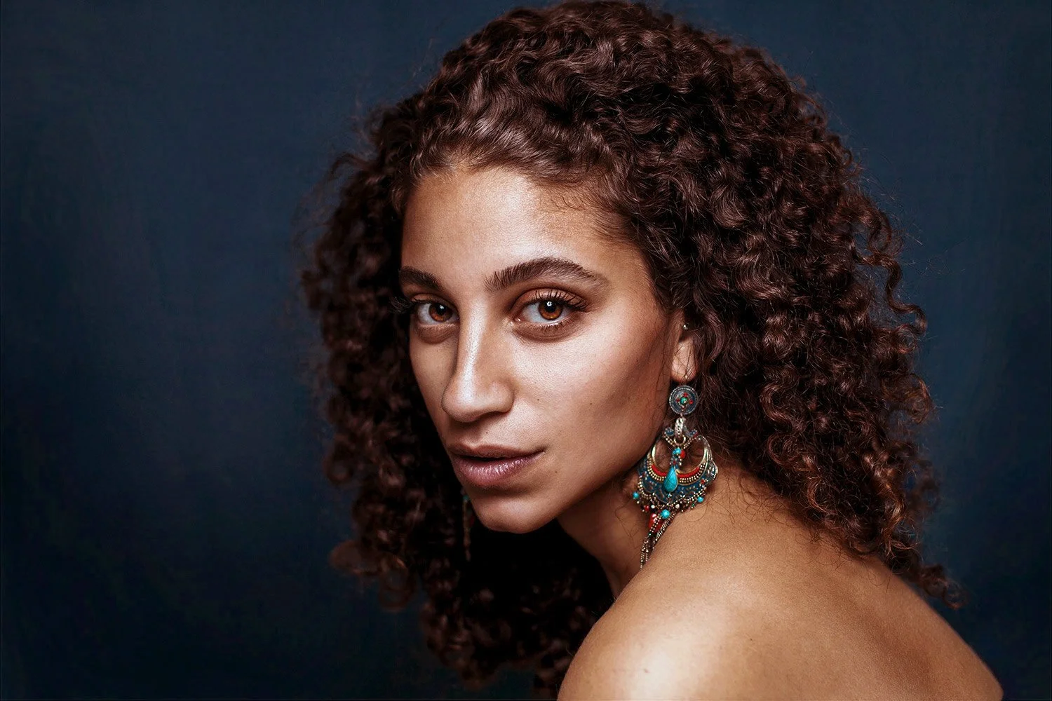 A woman with curly brown hair, wearing large colorful earrings, looking at the camera against a dark background.