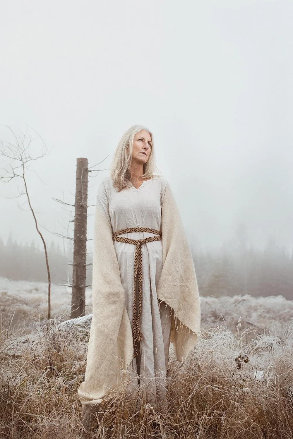 A woman with gray hair standing outdoors in a frosty, foggy field, wearing a beige robe and a woven belt, with leafless trees in the background.