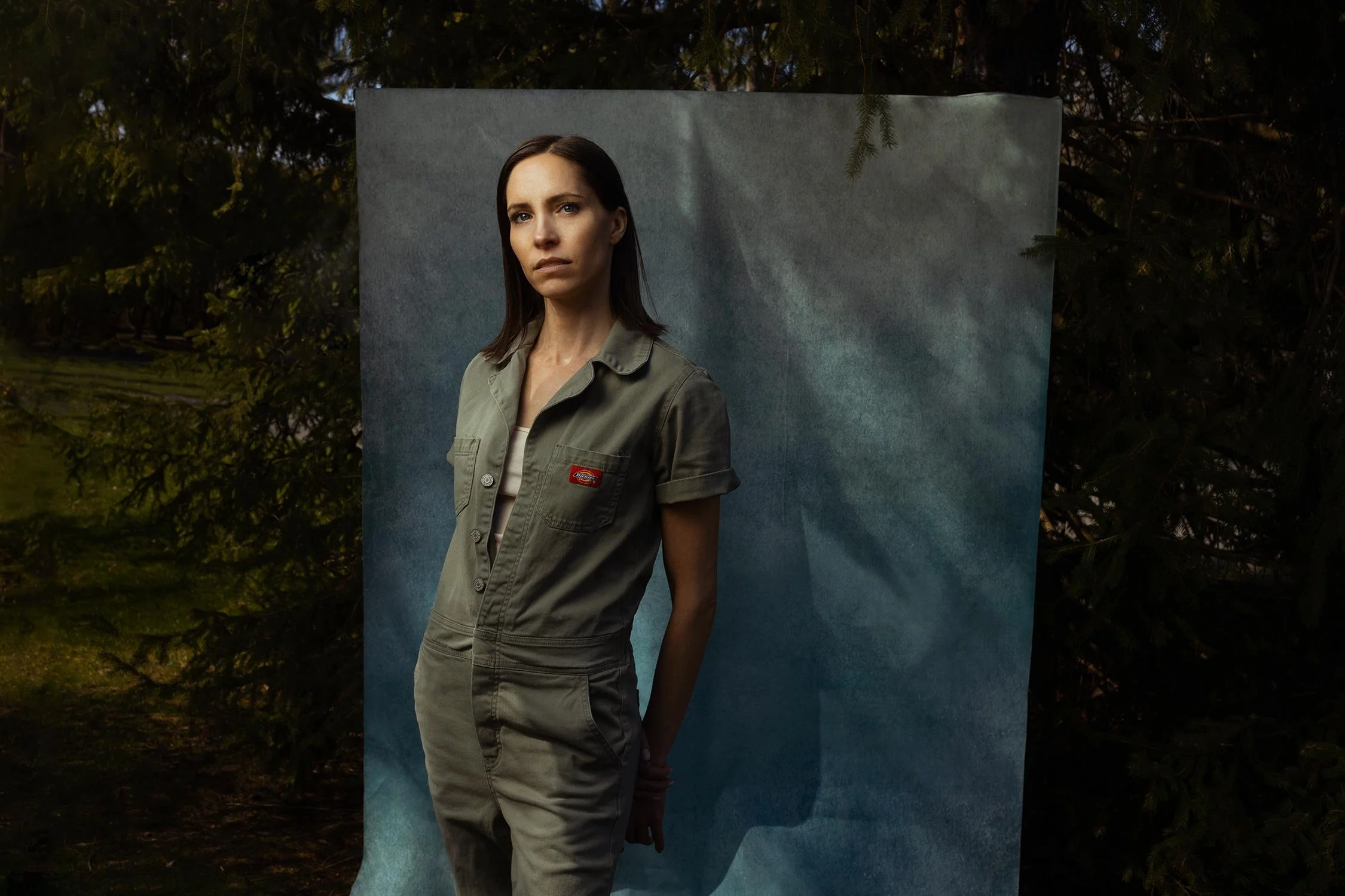 A woman wearing a green jumpsuit stands outdoors in front of a blue backdrop, with trees and greenery in the background.