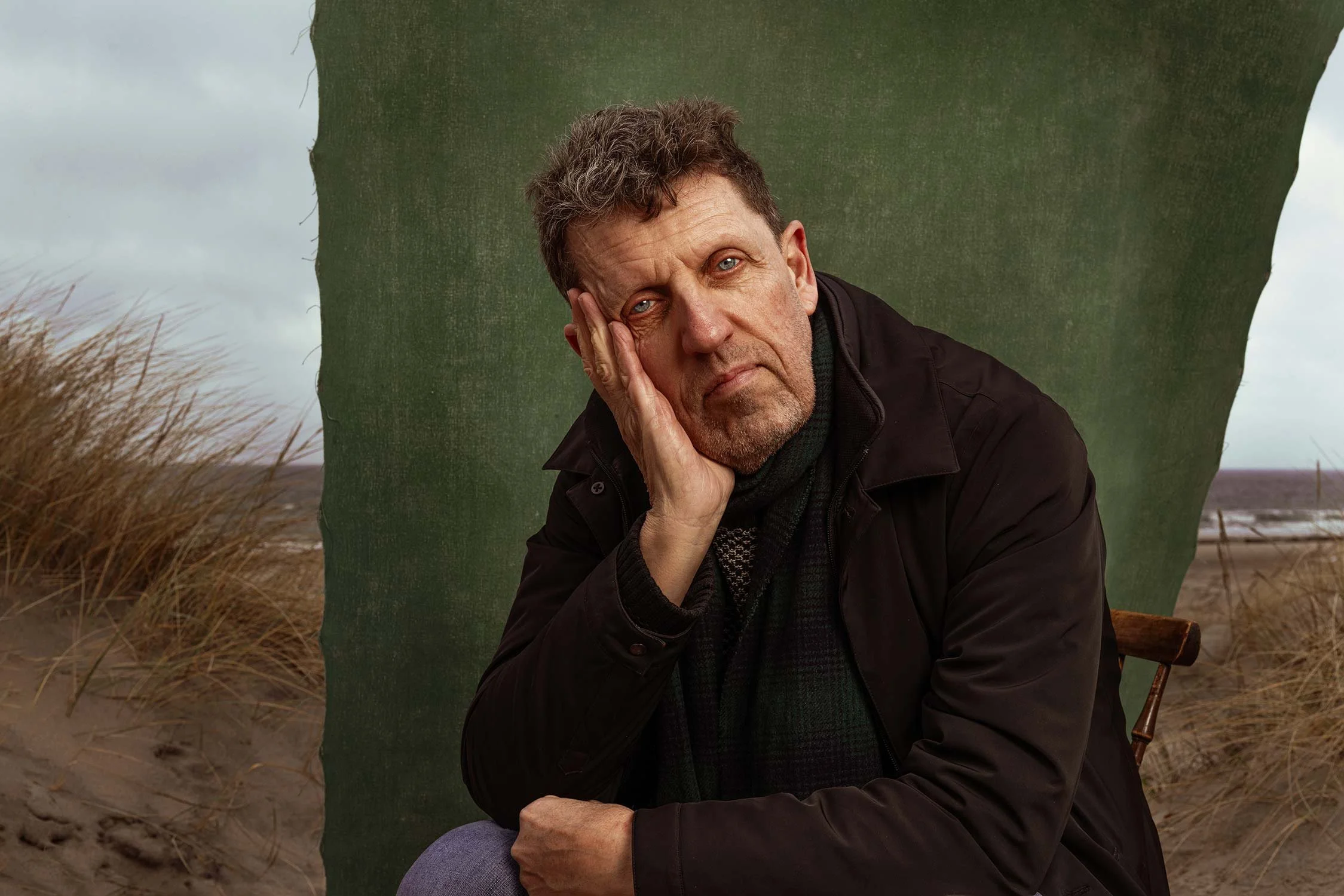 A middle-aged man with curly hair and a beard, resting his head on his hand with a pensive expression, sitting outdoors near dunes and tall grass, with a green textured backdrop.