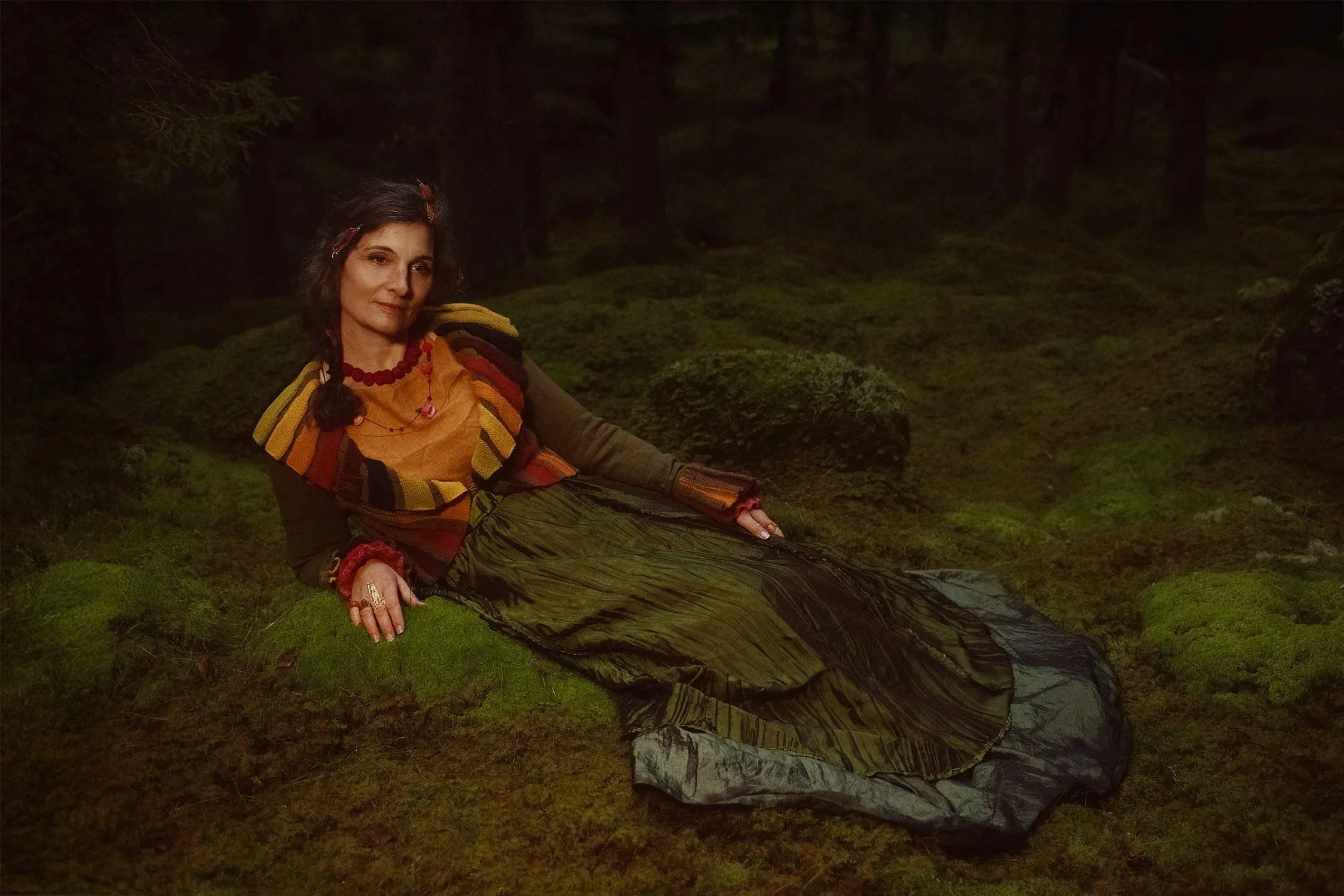 A woman with dark hair and colorful clothing lying on a mossy forest floor.