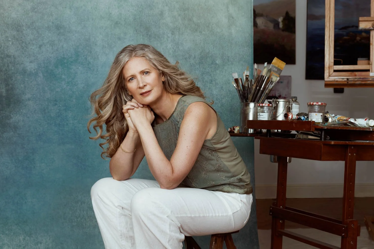 A woman with long wavy blonde hair, wearing a sleeveless gray top and white pants, sitting on a stool with her hands resting under her chin, in an art studio with paintbrushes and art supplies visible behind her.