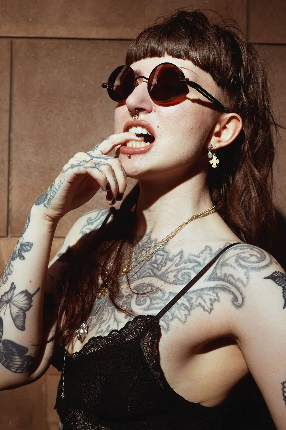 A tattooed woman with dark brown hair, wearing sunglasses, black lace top, earrings, necklaces, and biting her finger