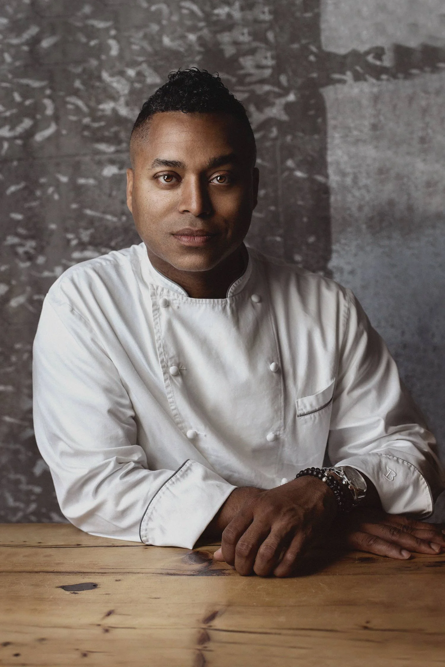 Portrait of a male chef in a white chef's coat sitting at a wooden table with a gray textured background.