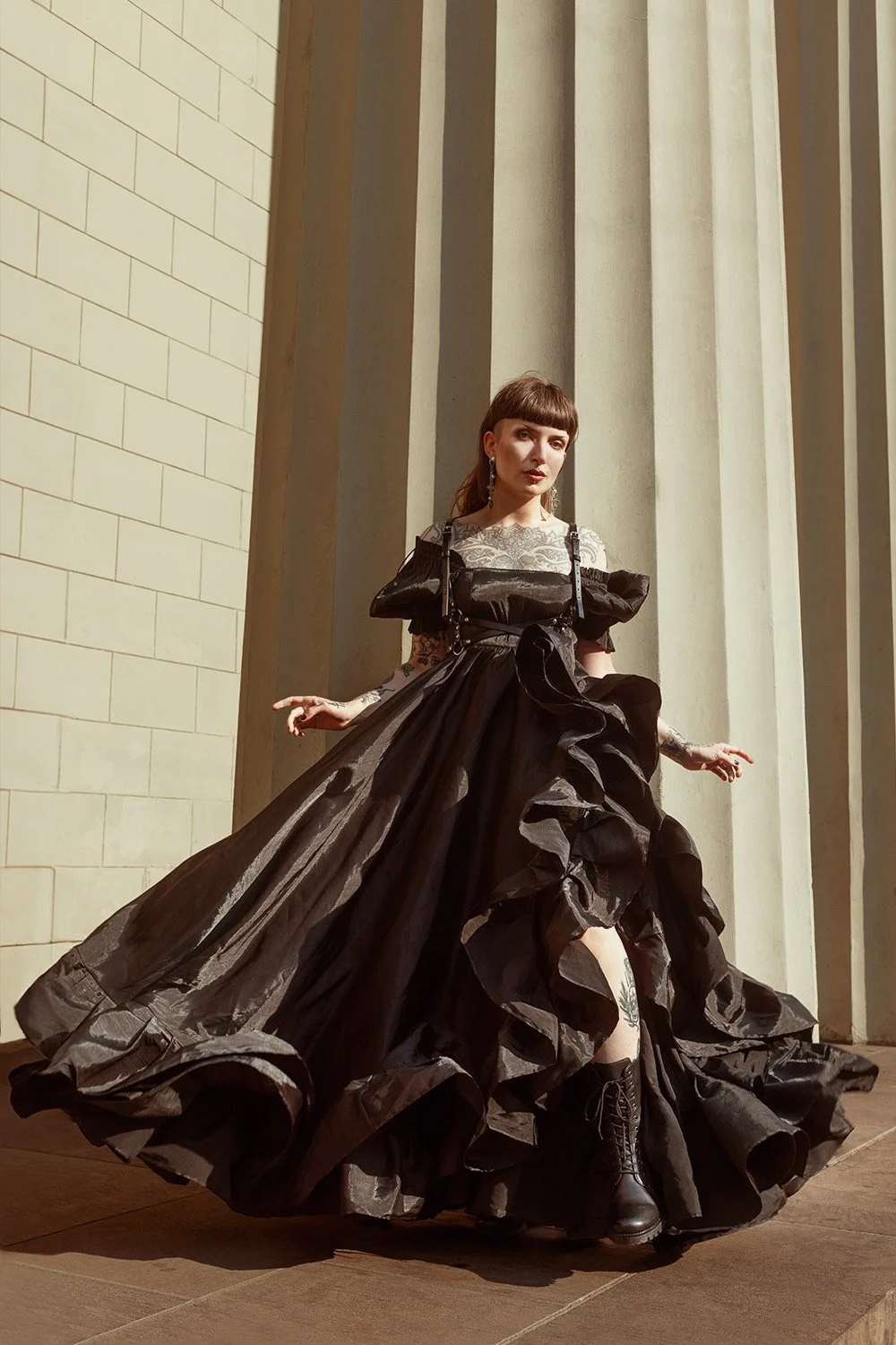 A woman wearing a gothic black dress with ruffles, standing against tall stone columns and a brick wall.