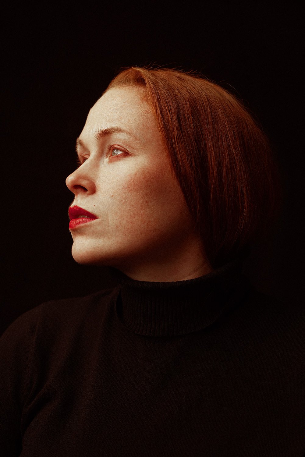 Side profile of a woman with red hair, fair skin, red lipstick, wearing a black turtleneck, against a black background.