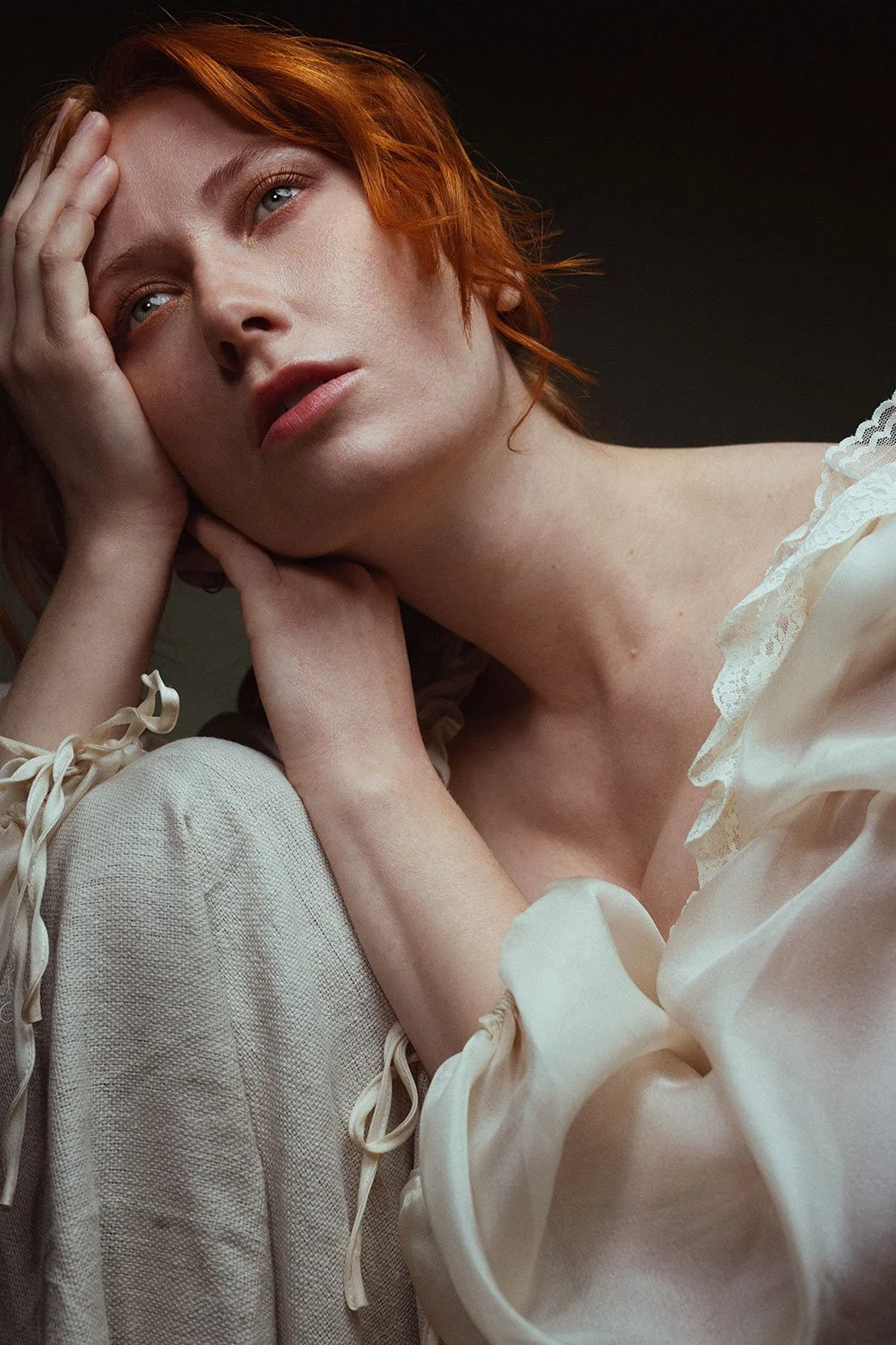 A woman with short red hair, wearing a cream-colored satin blouse with lace details, is resting her head on her hand with a contemplative expression, against a dark background.