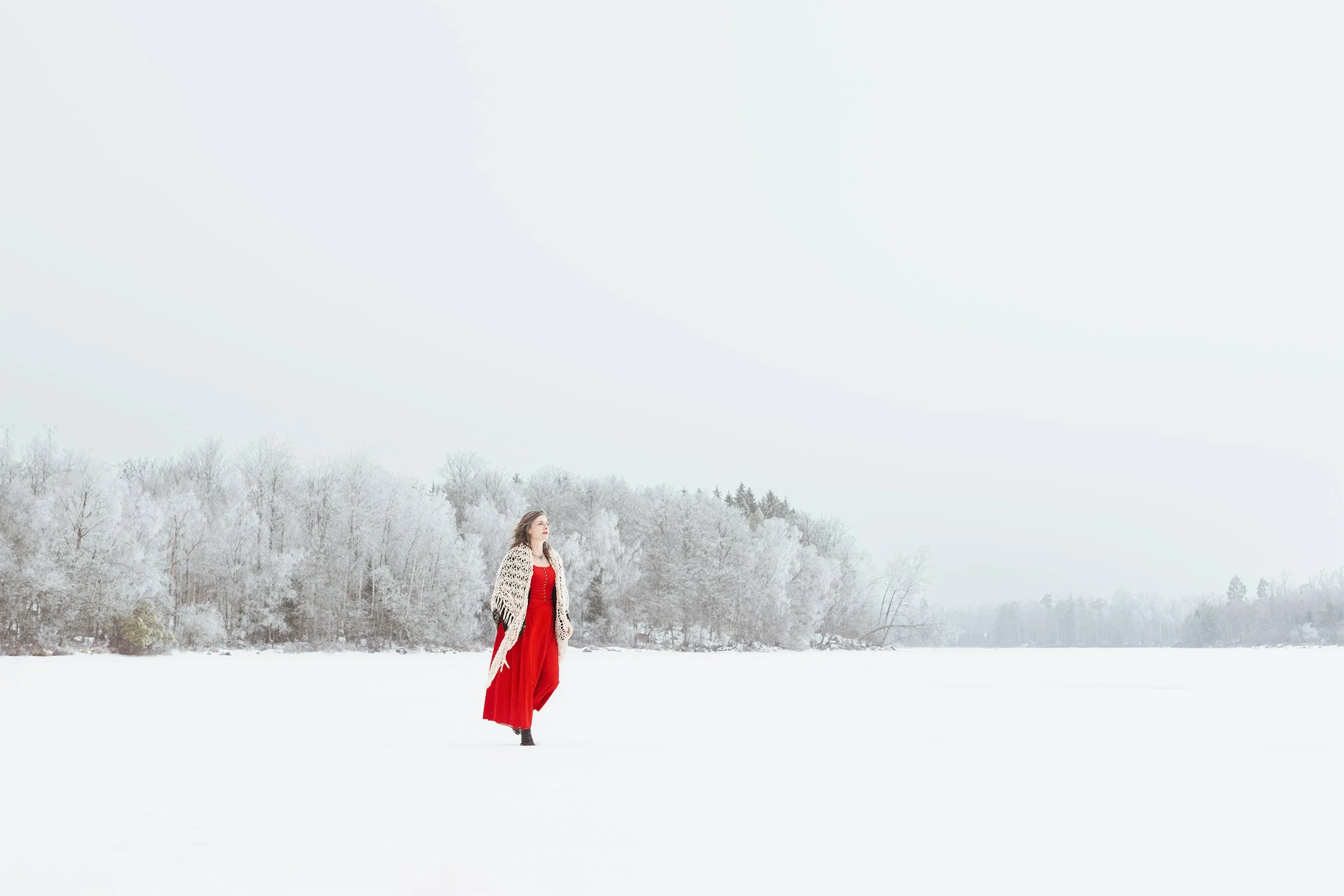 A woman in a red dress and a patterned shawl standing in a snowy landscape near a frozen lake with snow-covered trees in the background.