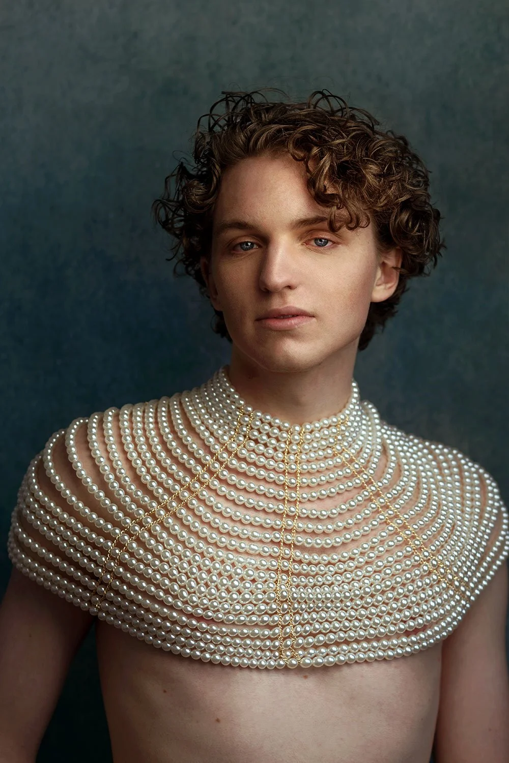 A young person with short curly hair wearing an elaborate pearl necklace draped over their shoulders and chest, against a dark background.