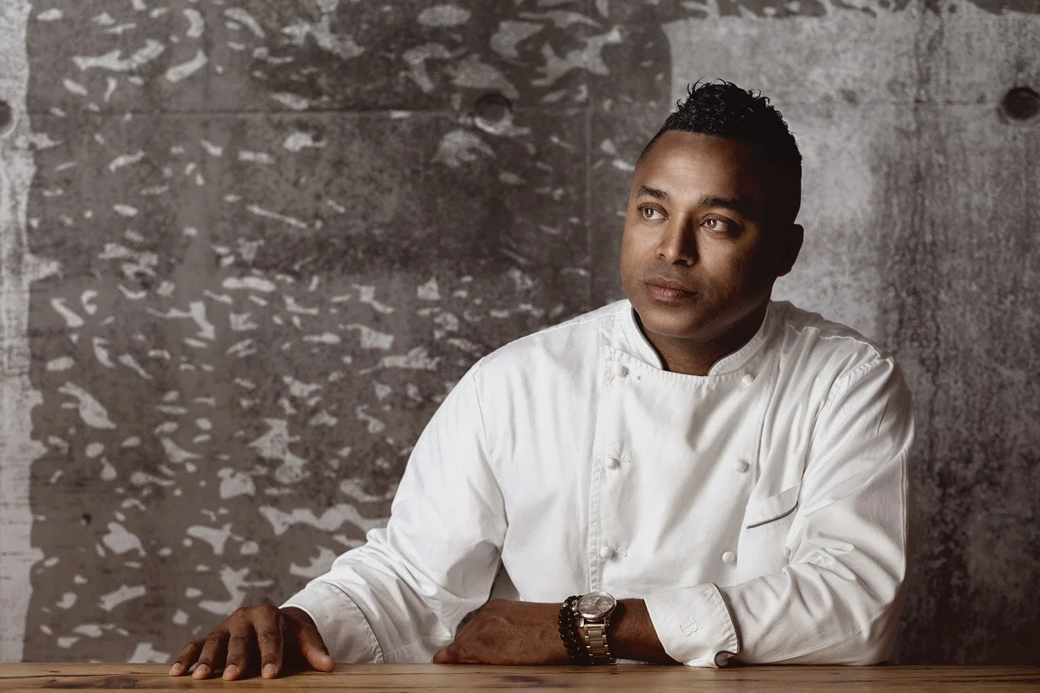 A man in a white chef's coat sitting at a wooden table, looking thoughtfully to the side, with a gray textured background.