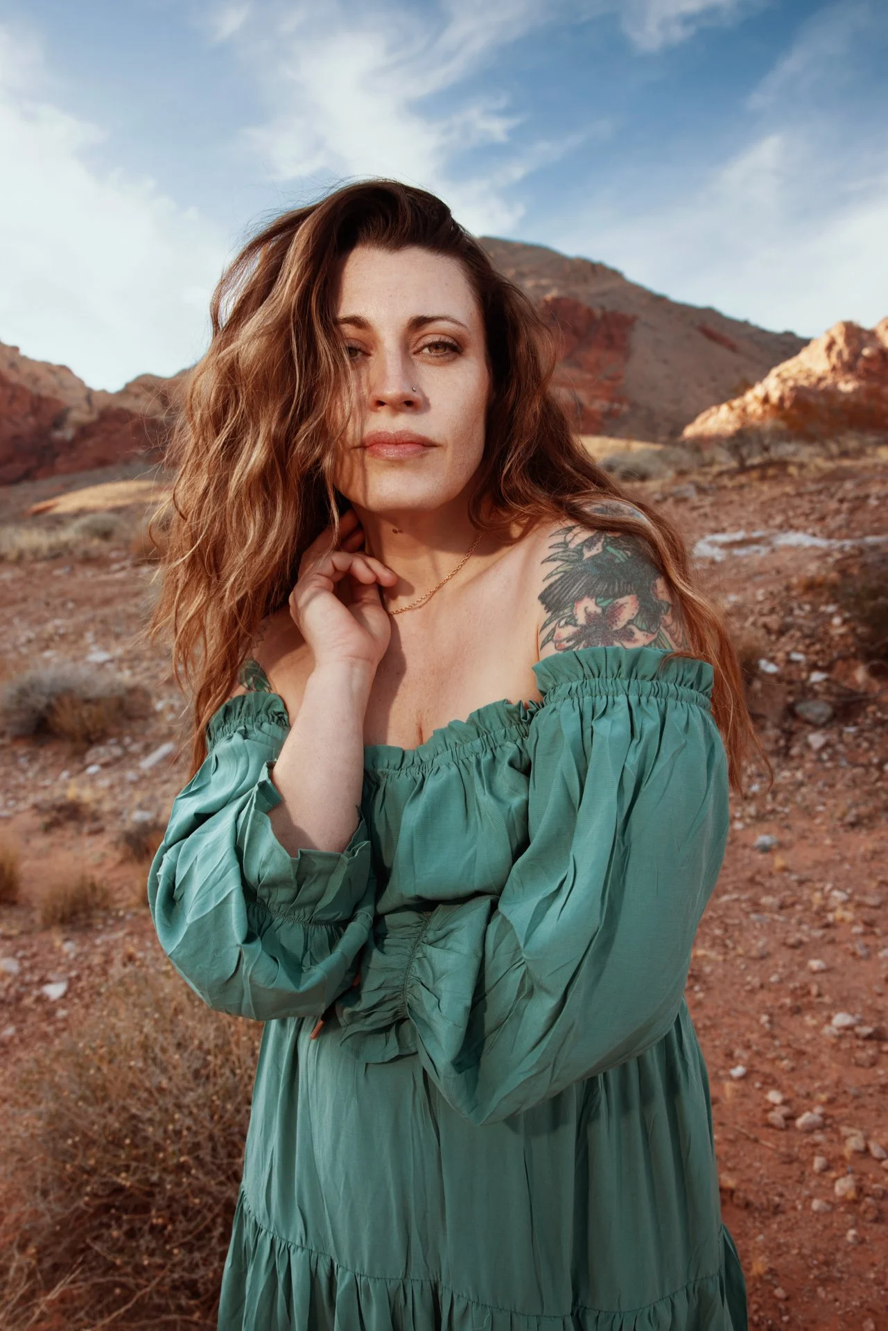 A woman with long wavy brown hair and a tattoo on her shoulder, wearing a green off-shoulder dress, standing outdoors with a rocky mountainous landscape in the background.