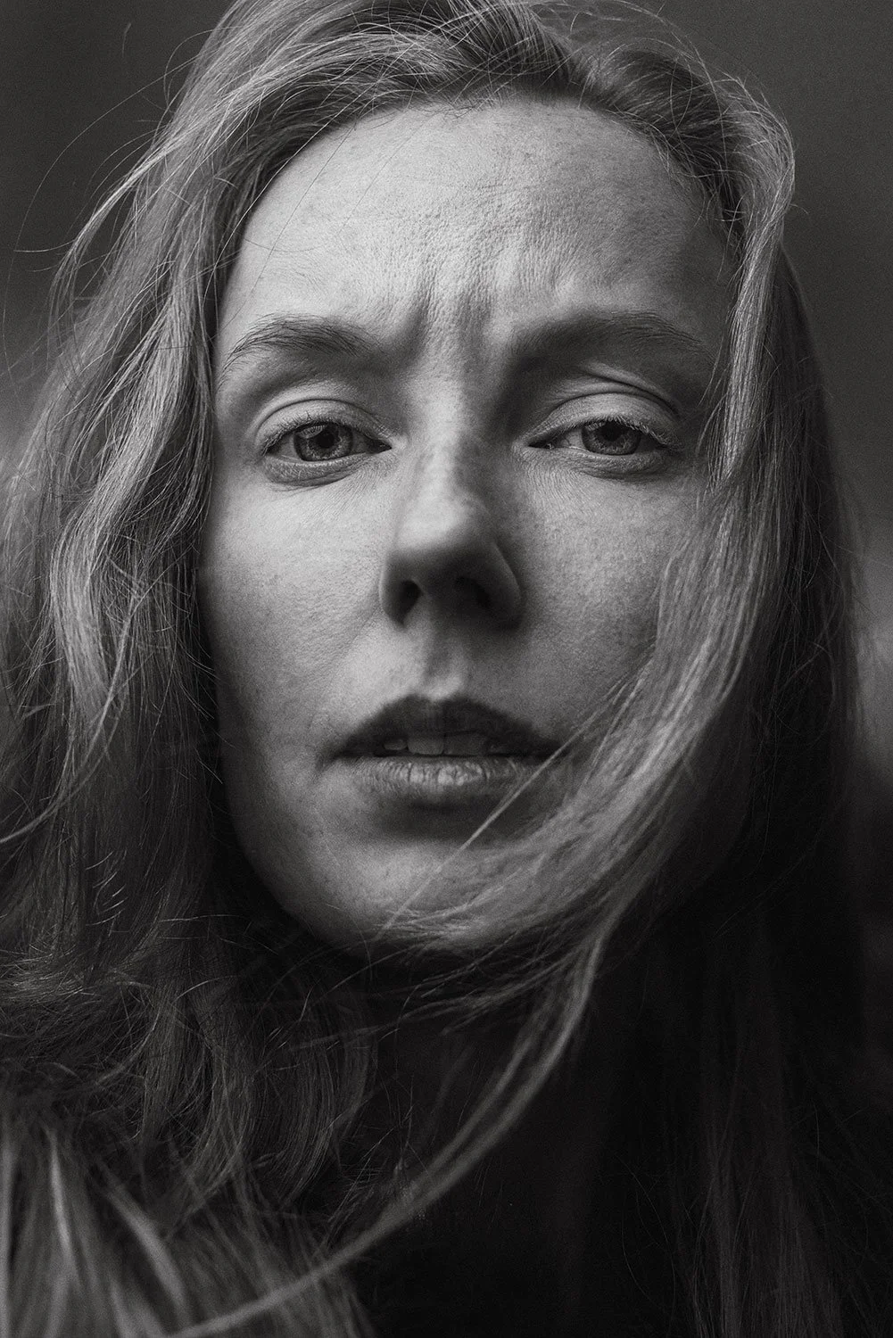Close-up black and white portrait of a woman with long hair, with windblown strands across her face and a serious expression.