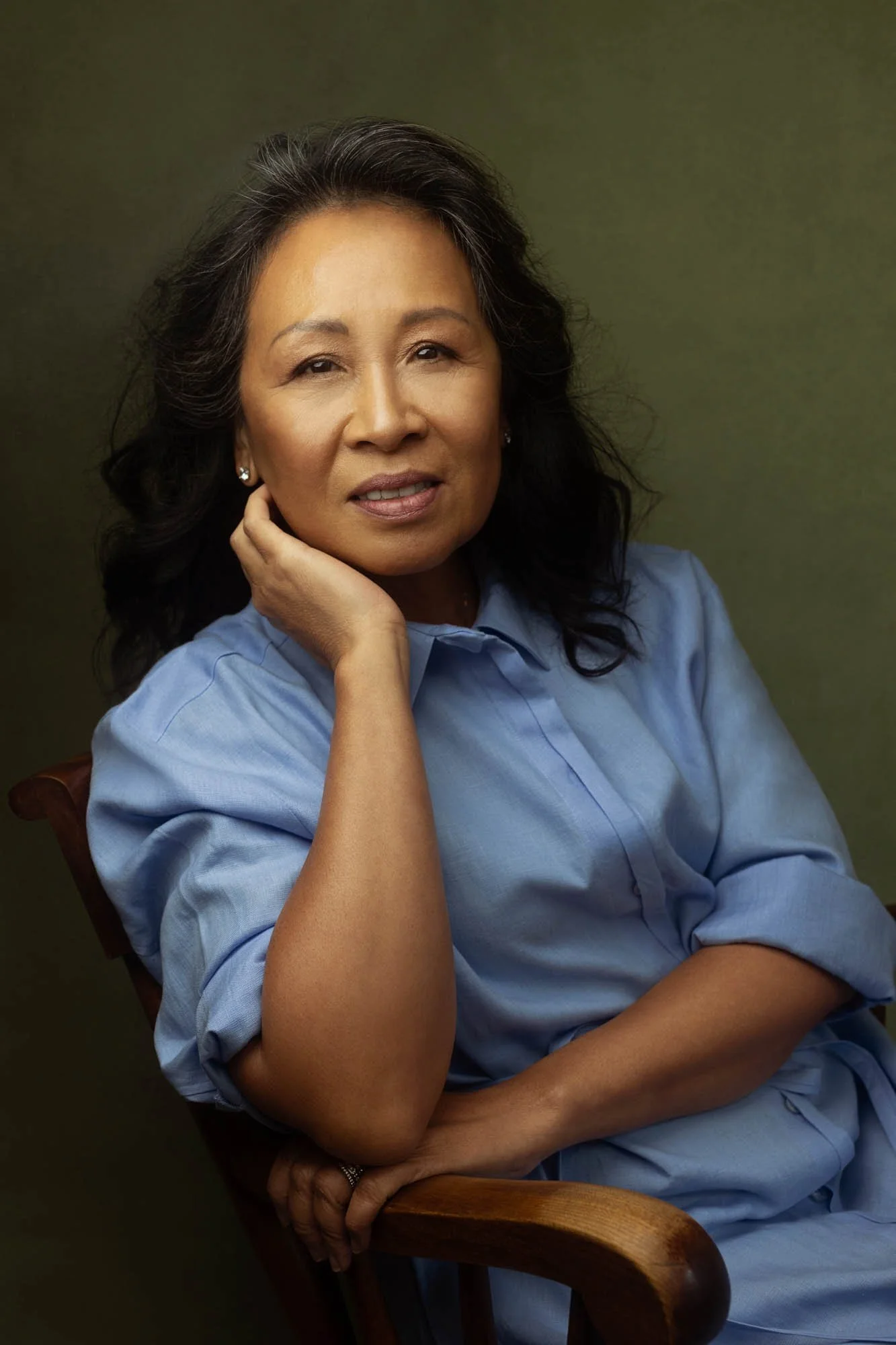 Portrait of an older, Asian woman with long black hair, wearing a light blue shirt, sitting on a wooden chair, holding her face with one hand, against a dark green background.