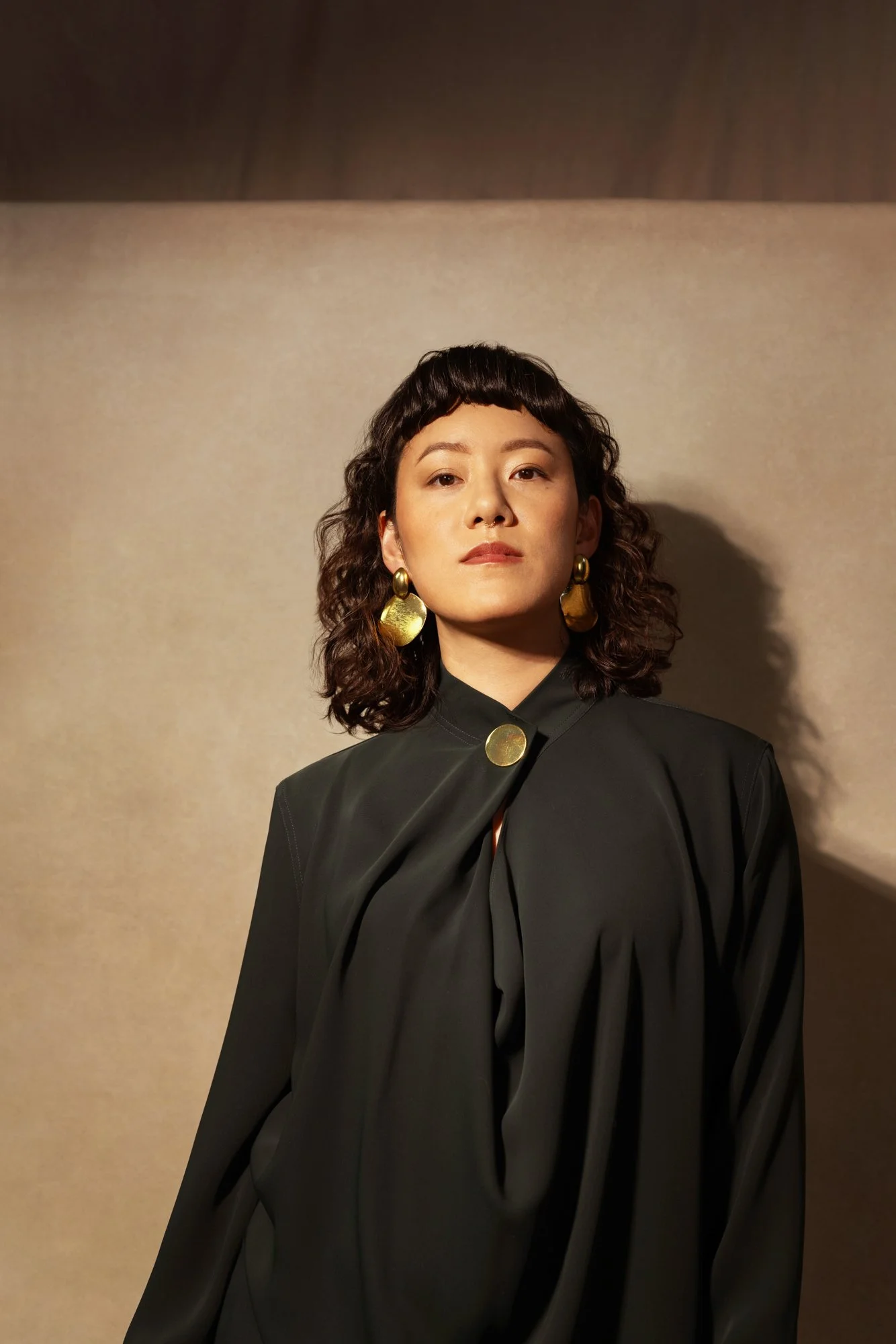 A woman with curly dark hair and gold jewelry, wearing a black outfit, looking confidently at the camera against a beige backdrop.