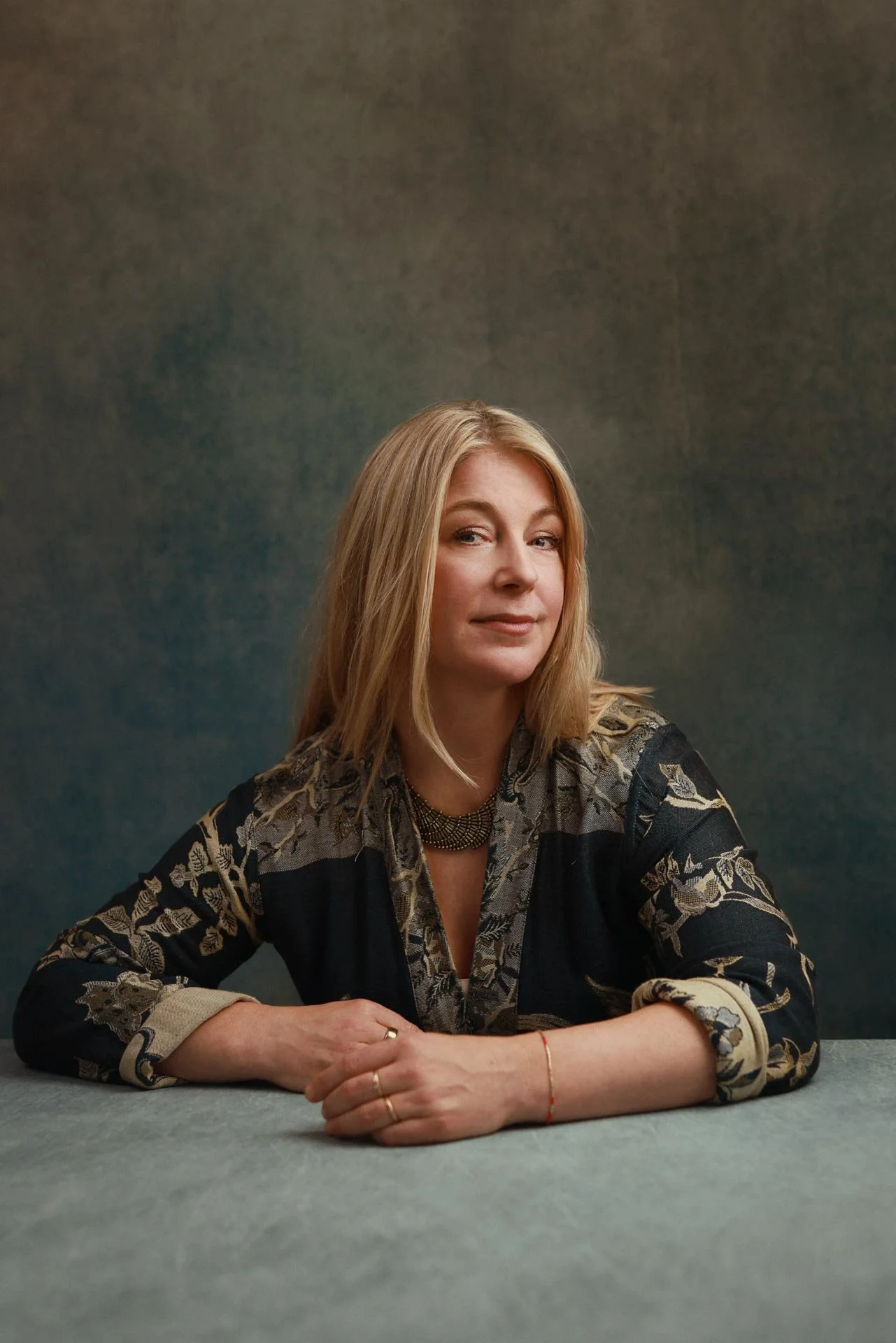 A woman with blonde hair, wearing a patterned dark kimono, sits at a table against a textured dark, blue, background.