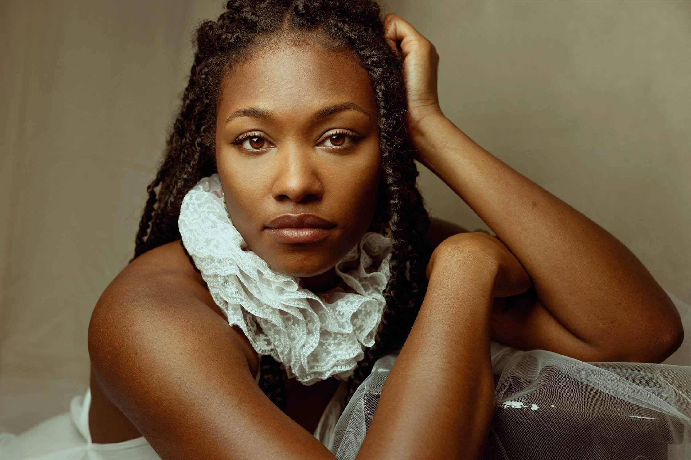 A woman with dark skin, with braids, looking directly at the camera, resting her chin on her hand, wearing a ruffled white lace collar.
