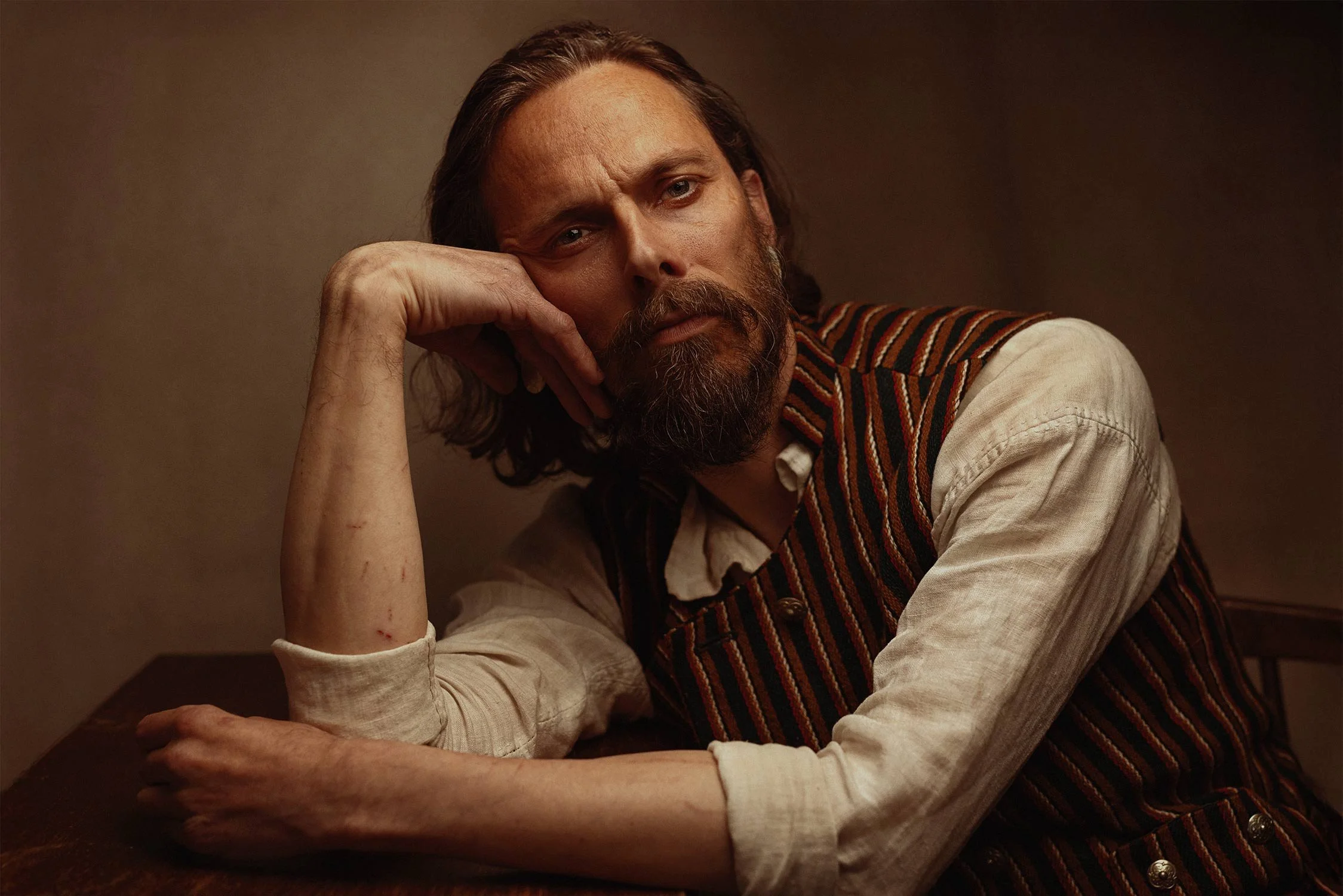 A man with long hair and a beard, wearing a beige shirt and a striped vest, sits at a table with a serious expression, resting his head on his hand.