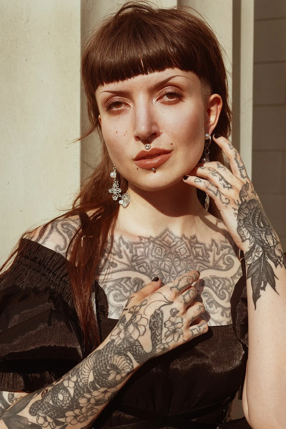 A woman with red hair, tattoos on her chest and arms, wearing earrings and jewelry, posing outdoors in sunlight.