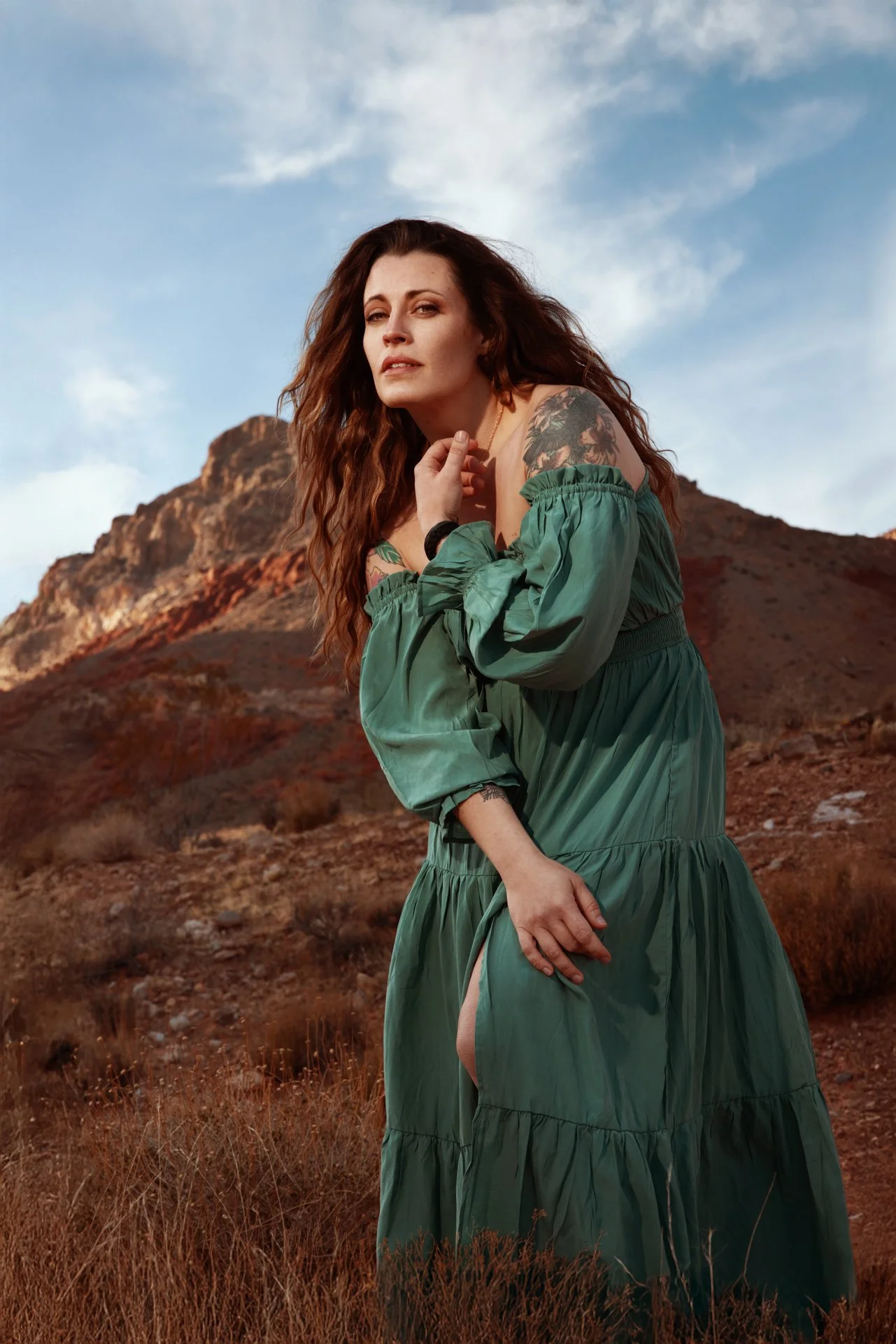 A woman with long, curly red hair and tattoos on her shoulder and arm, wearing a green off-shoulder dress, standing outdoors in front of a rocky, desert-like hill under a partly cloudy sky.