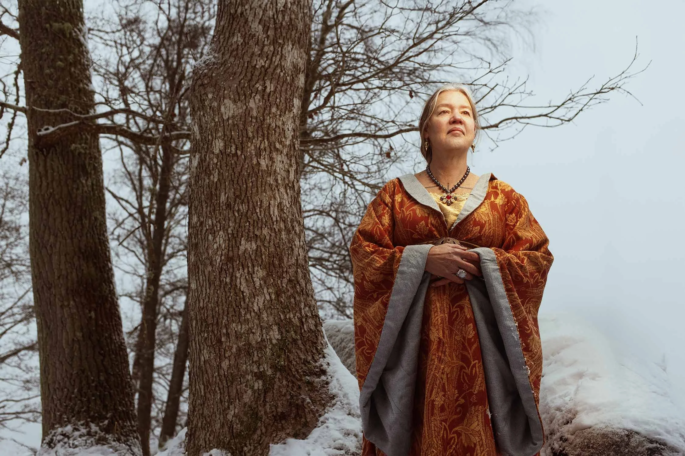 A woman standing outdoors on a snowy day, wearing an ornate, reddish-brown and gray cape, with trees in the background.