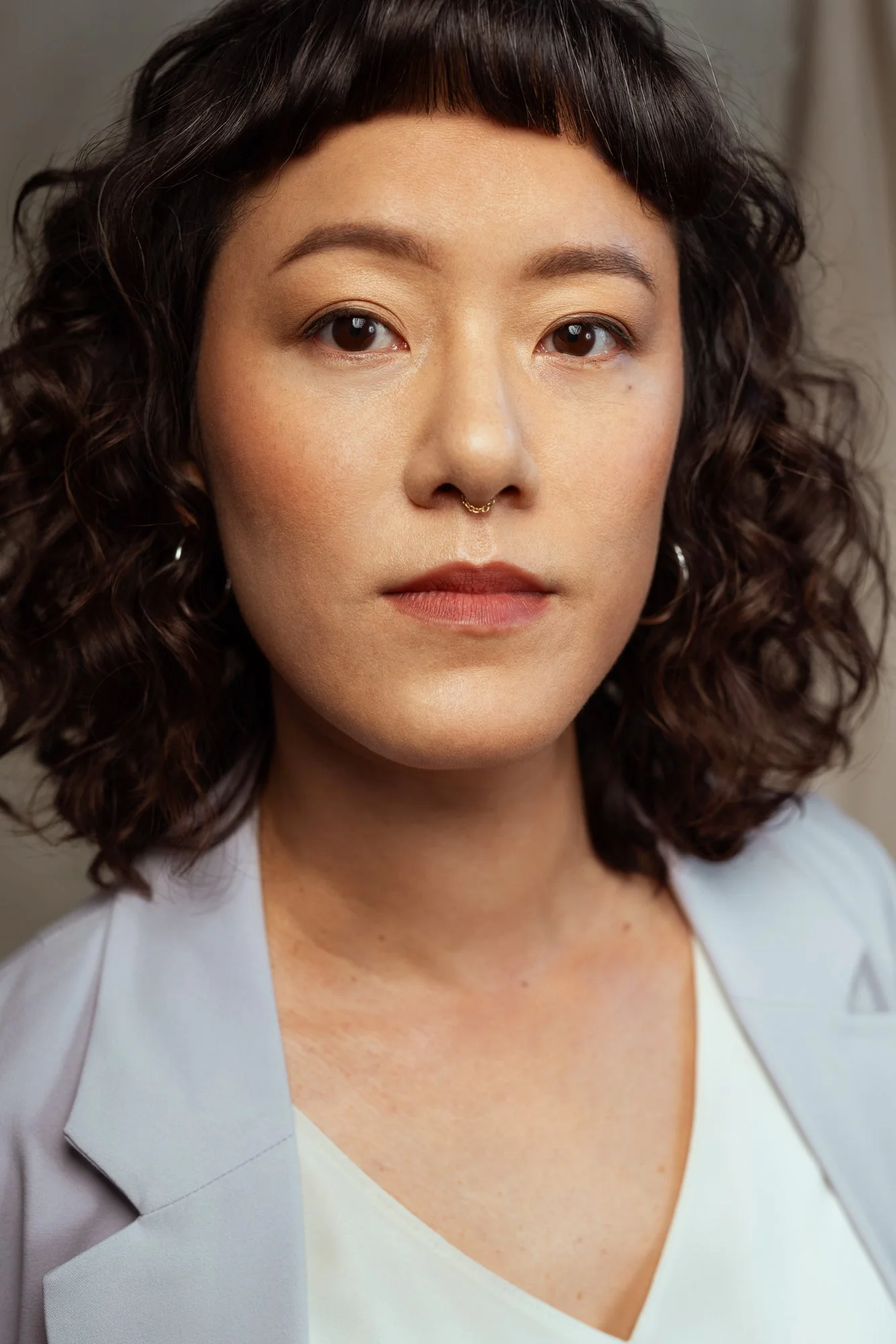Close-up portrait of an Asian woman with curly dark hair and a septum piercing, wearing a light lavender blazer and white top, looking directly into the camera with a neutral expression.