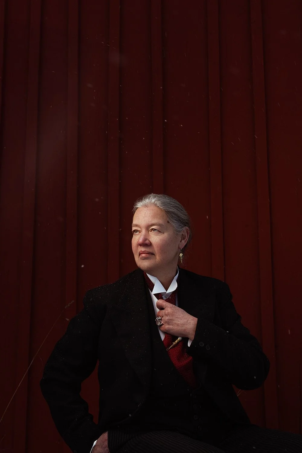A woman with gray hair wearing a black suit and white shirt, sitting in front of a dark wooden wall, looking thoughtfully to her right.