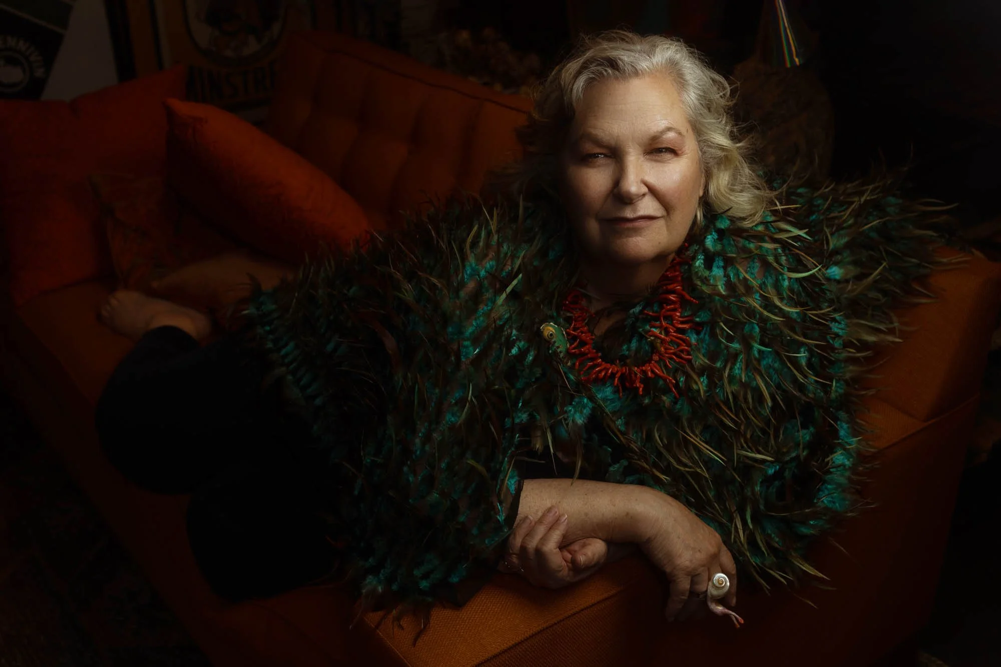 An older woman with gray hair sitting on an orange couch, wearing a feathered garment and a red coral necklace, looking at the camera.