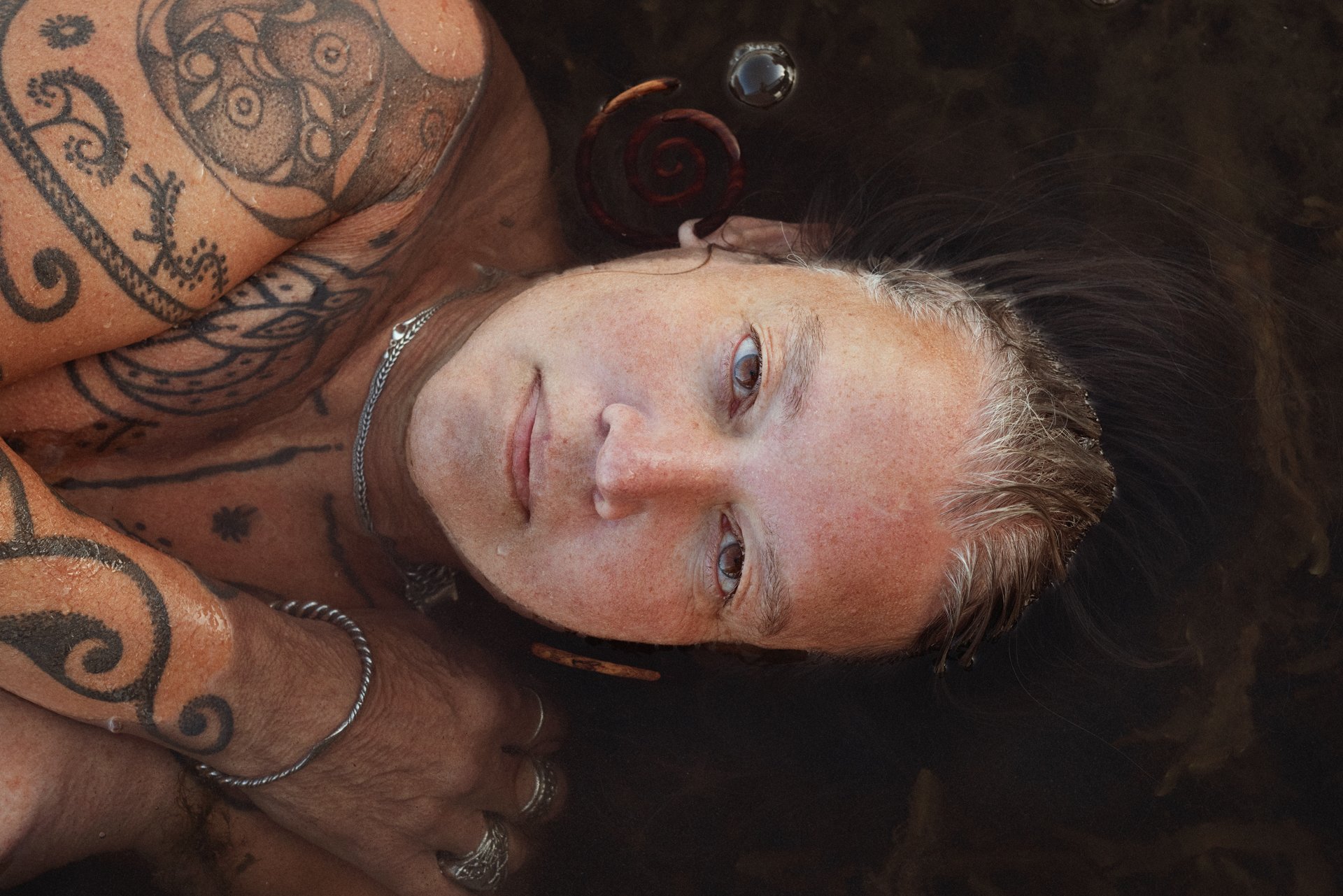 A person with long, dark hair lying in water, with visible tattoos on their arms and chest, smiling slightly, wearing rings and necklaces.