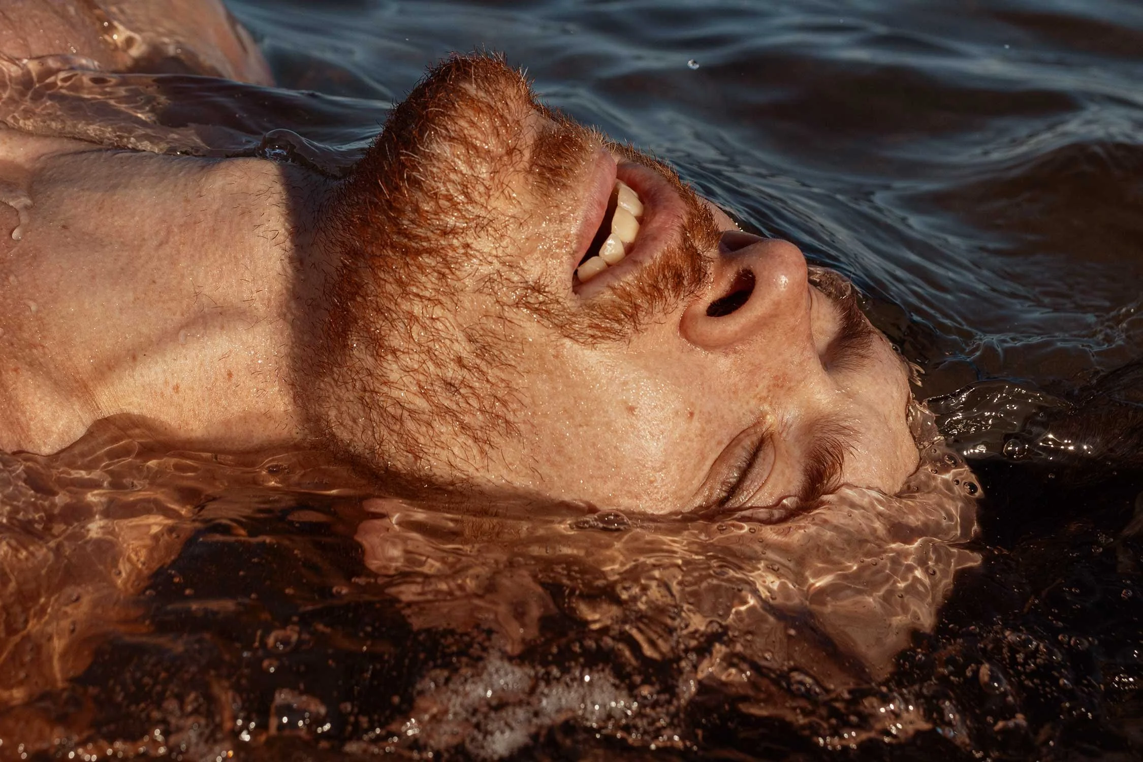 Man with red hair and beard smiling and floating in water with eyes closed.