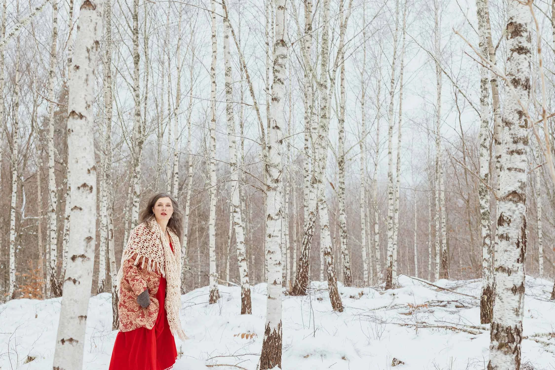 A woman with long hair wearing a red dress and a beige shawl stands in a snow-covered forest of birch trees, looking to the side.