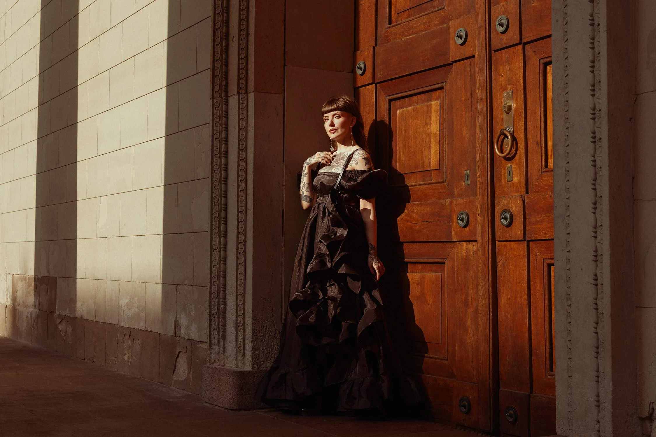 A woman in a black ruffled gown standing against a large wooden door, illuminated by warm sunlight, with her hand on her chest and looking at the camera.
