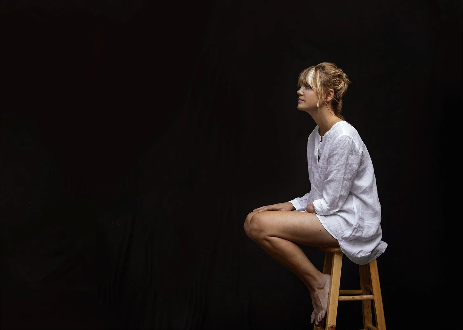 A one-armed woman with blonde hair sitting on a wooden stool against a black background, wearing a white long-sleeve shirt and looking to the side.
