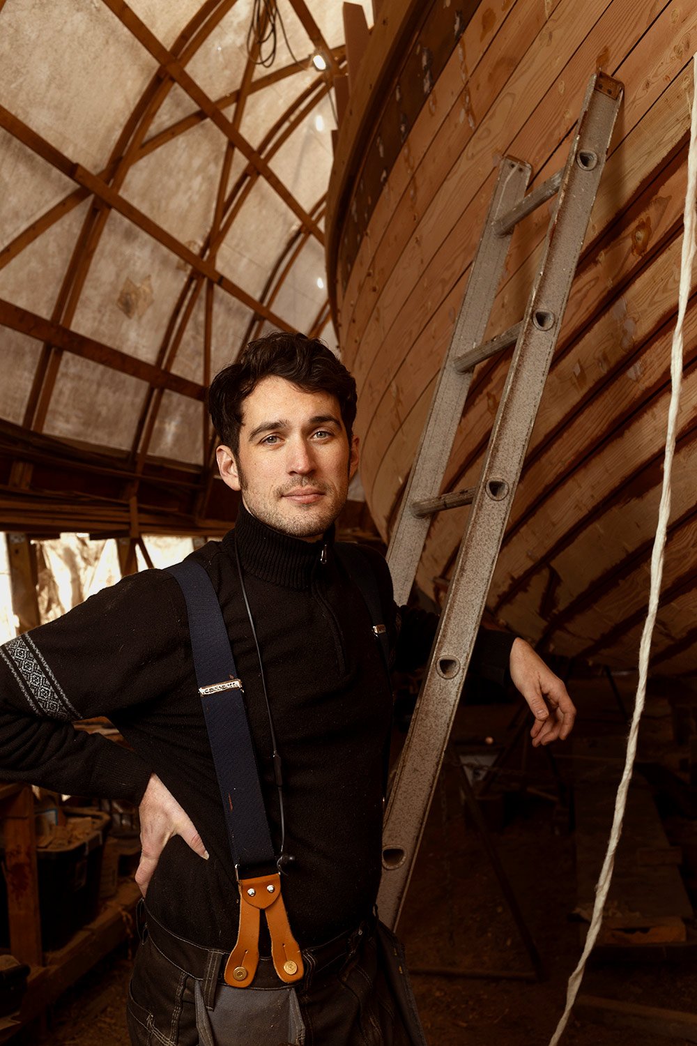 A man poses next to a ladder inside a wooden boat under construction, with the boat's curved wooden frame behind him.