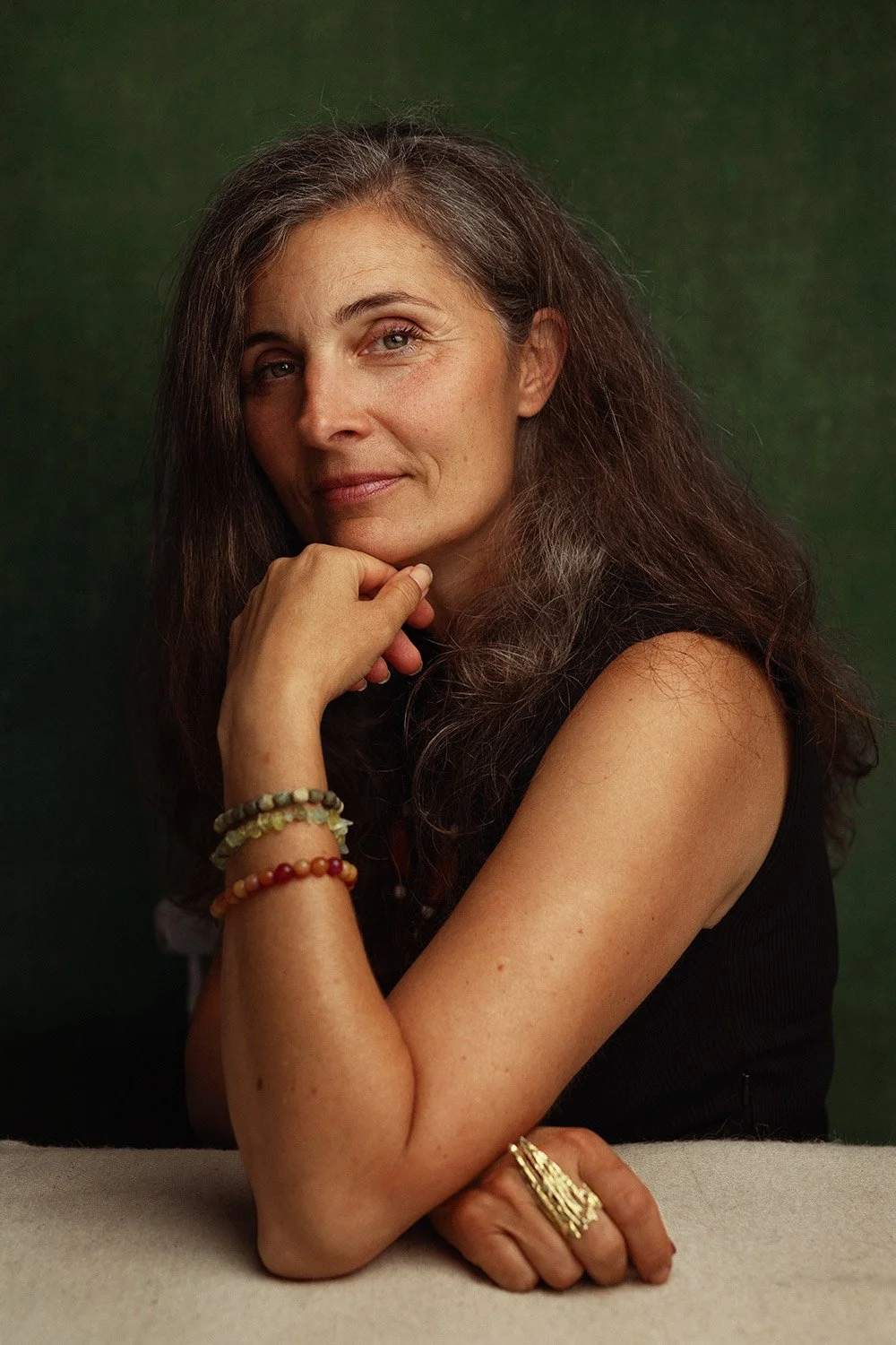 A woman with long, wavy brown hair and fair skin resting her chin on her hand, wearing a black sleeveless top, multiple colorful beaded bracelets, and rings, against a dark green background.
