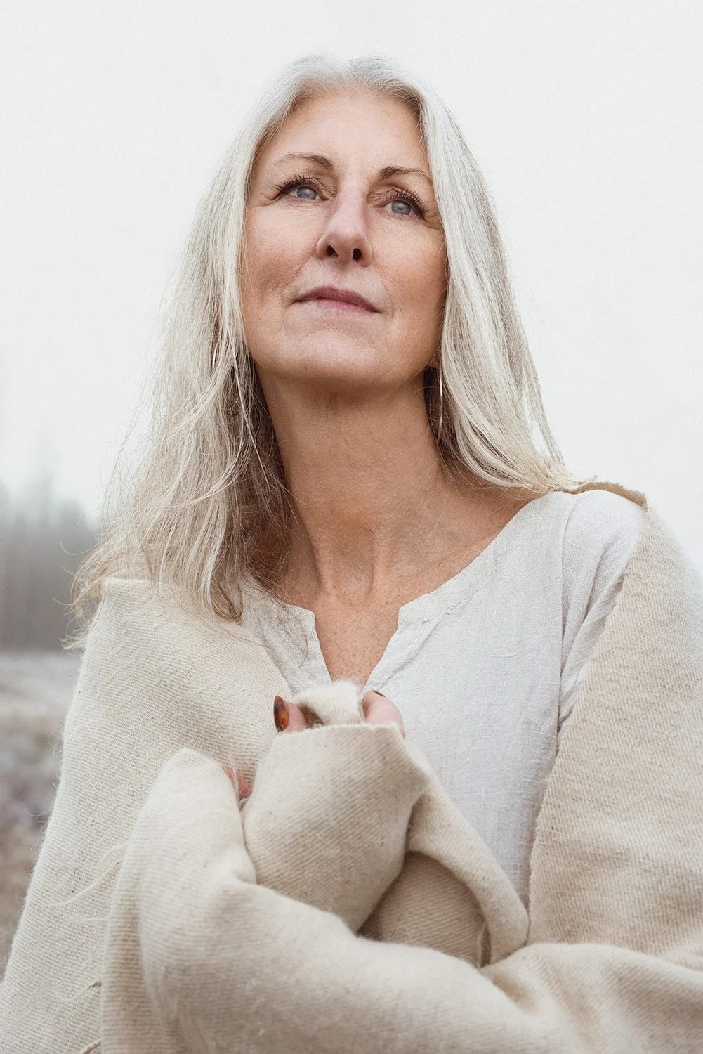 A mature woman with long gray hair wrapped in a beige blanket outdoors.