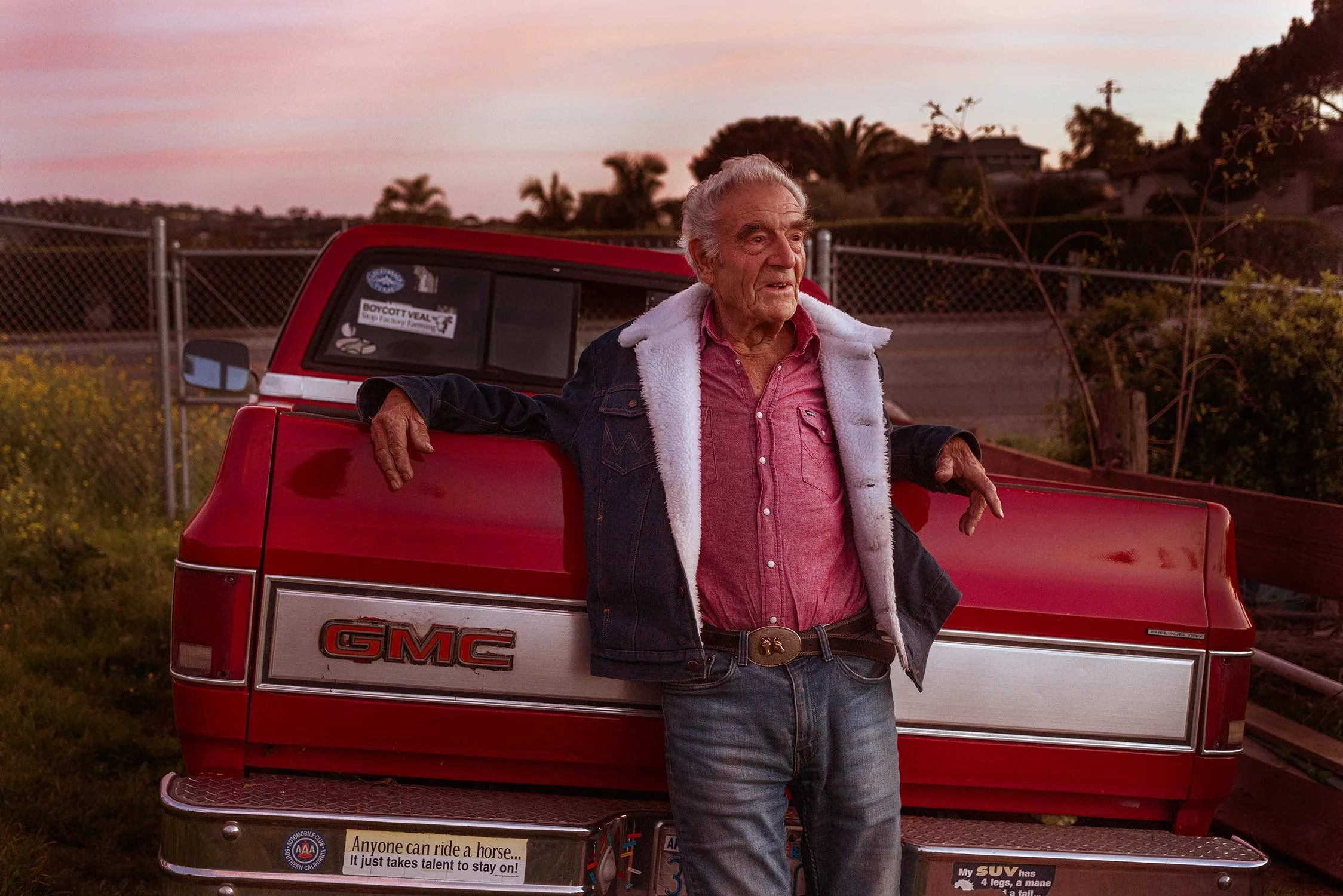 An elderly man leaning against the back of a red GMC pickup truck at sunset, wearing a pink button-up shirt, jeans, and a denim jacket with a white fleece lining, with trees and a fence in the background.