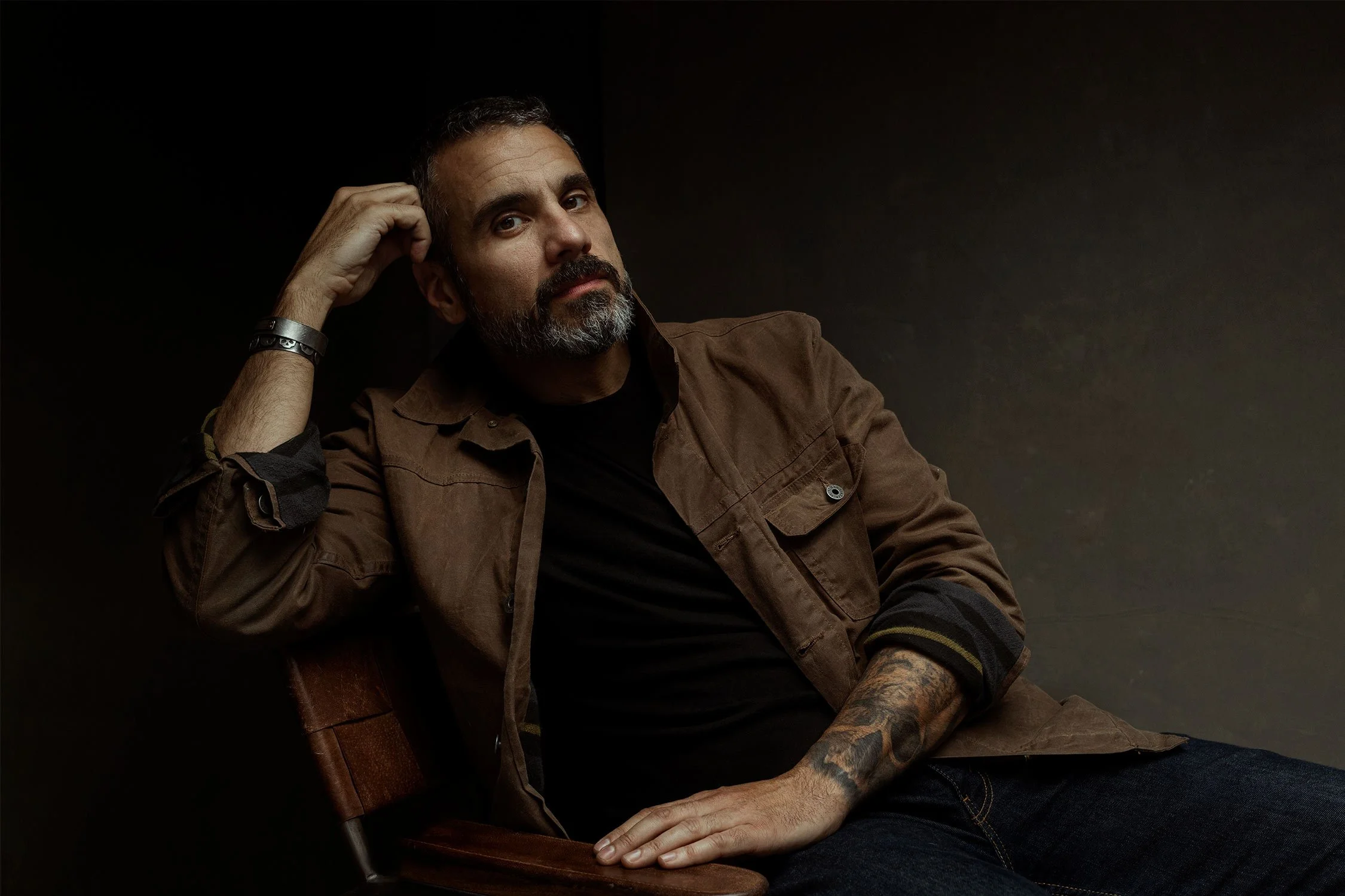 A man with a beard and tattoos, wearing a brown jacket and black shirt, sitting on a wooden chair against a dark background, looking into the camera with his hand behind his head.