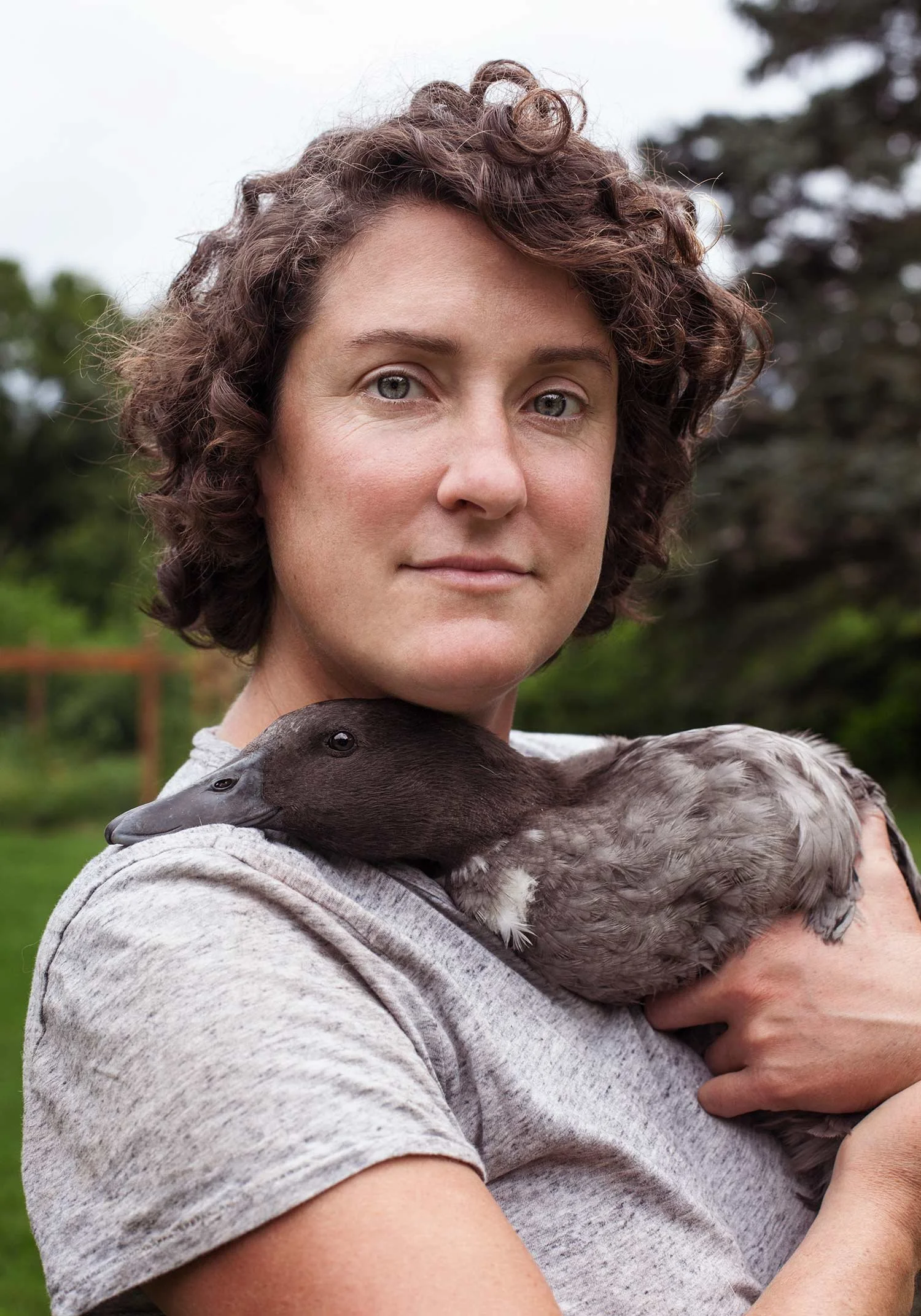 A woman holding a duck on her shoulder outdoors, with a background of green trees and cloudy sky.