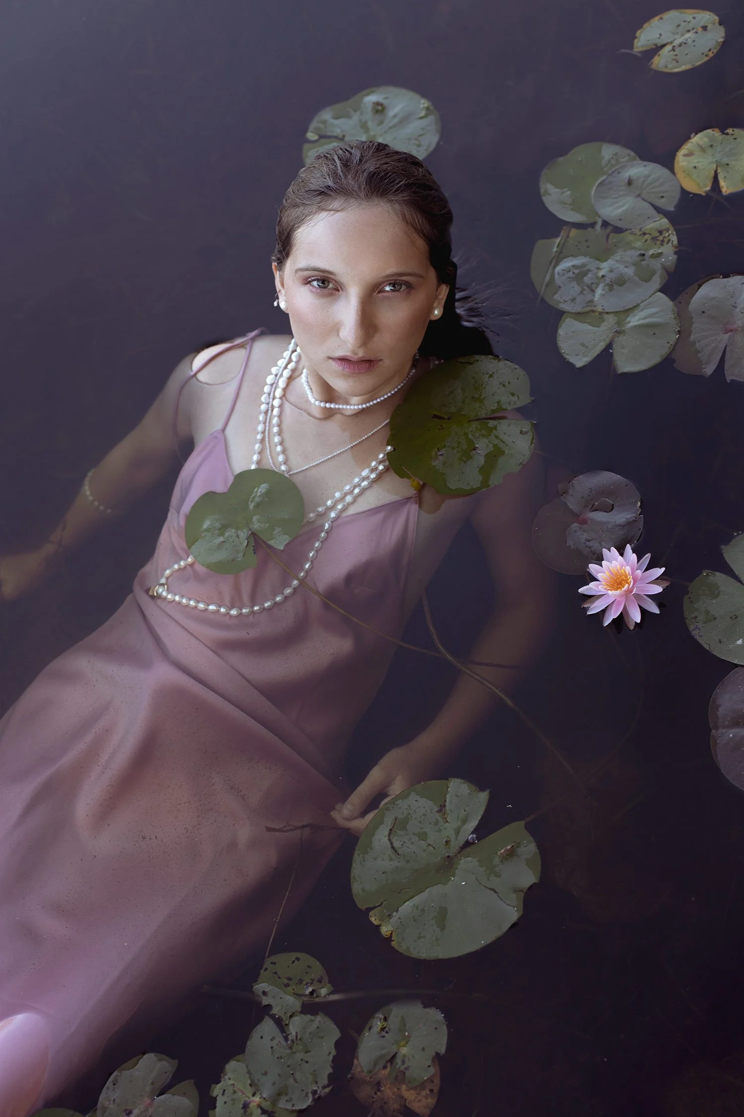 A woman in a pink satin dress and pearl jewelry floating in a pond with lily pads and a pink water lily.
