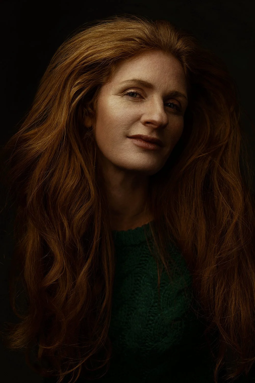 Close-up portrait of a woman with long, wavy red hair, wearing a dark green sweater, against a black background.