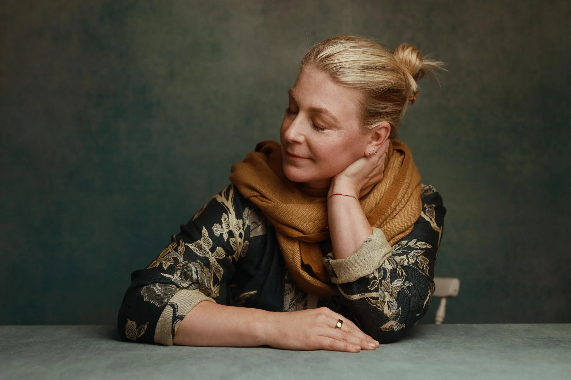 A woman with blonde hair tied back, wearing a patterned black and beige top and a brown scarf, resting her head on her hand with her eyes closed, sitting at a table against a dark green background.