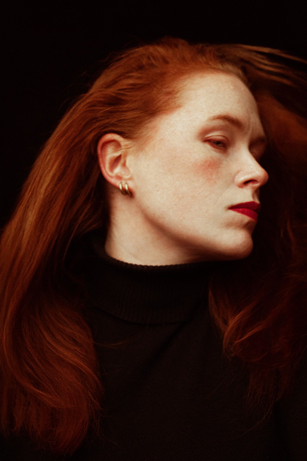 Profile of a woman with long red hair, wearing a black turtleneck, red lipstick, and gold hoop earrings against a dark background.