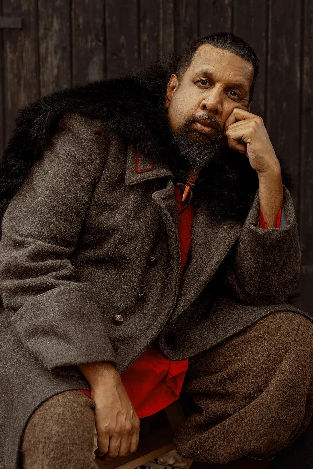 A man with long dark hair, a beard, and mustache sitting in front of a dark wooden background. He is wearing a gray coat with a black fur collar, and red clothing underneath. He is resting his head on his right hand with his elbow on his knee, lookin