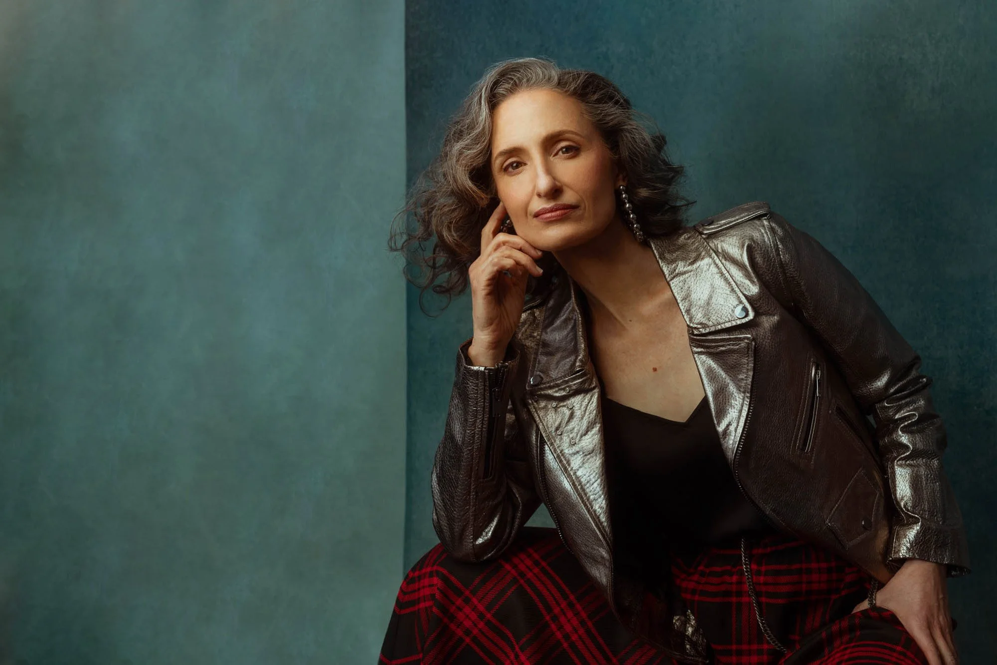 A woman with curly gray hair wearing a black top, a metallic jacket, and red plaid pants, sitting against a teal background.