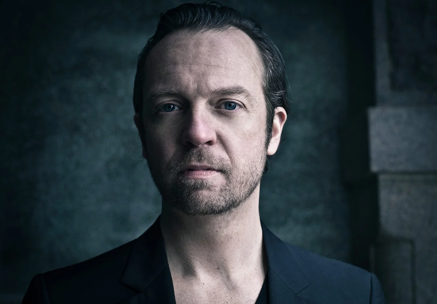 Close-up portrait of a man with short dark hair, light skin, blue eyes, and a beard, wearing a dark jacket against a dark, textured background.