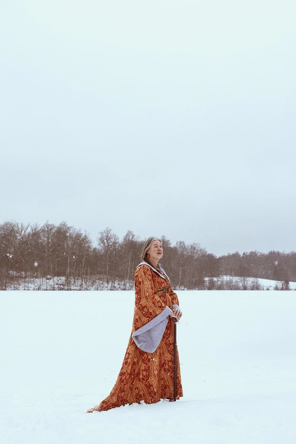 A woman dressed in an elaborate, ornate orange and gold gown with grey accents, standing outdoors in a snow-covered landscape with a background of leafless trees under a cloudy sky.