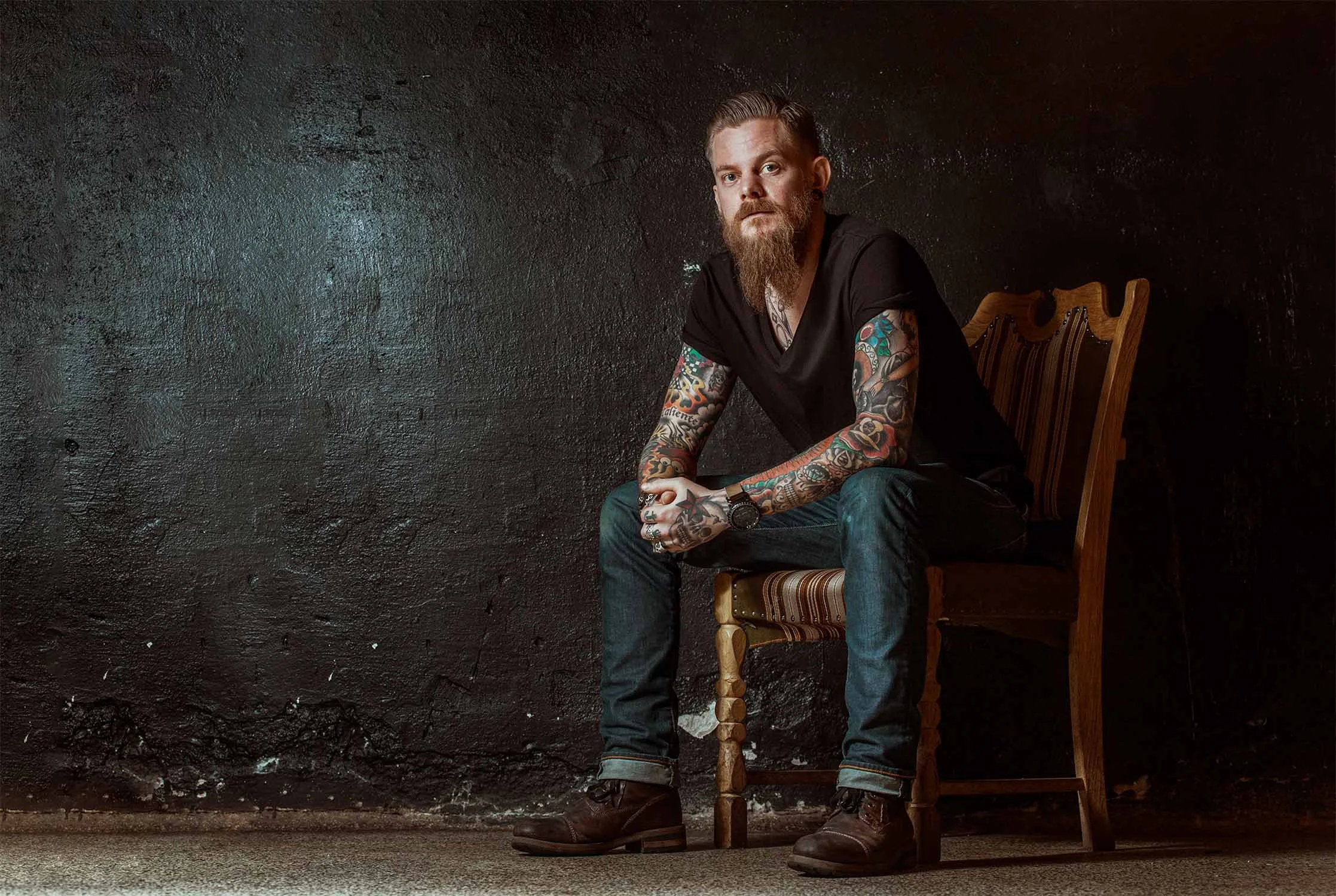 A man with a beard and tattoos sitting on a wooden chair against a black textured wall, wearing a black t-shirt, jeans, and leather boots.