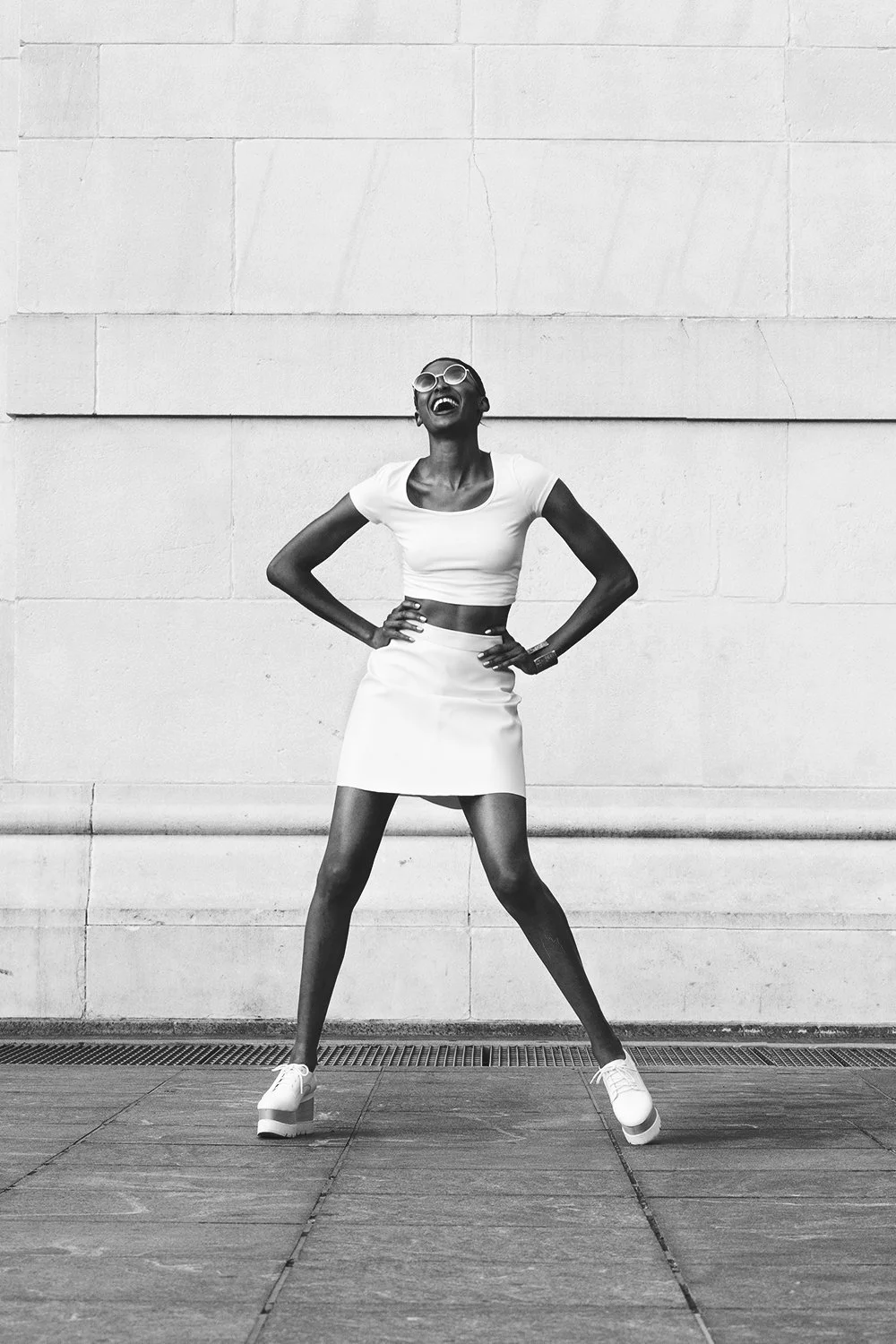 A woman in a white crop top and skirt wearing sunglasses and platform shoes, laughing with her hands on her hips, standing against a textured wall.